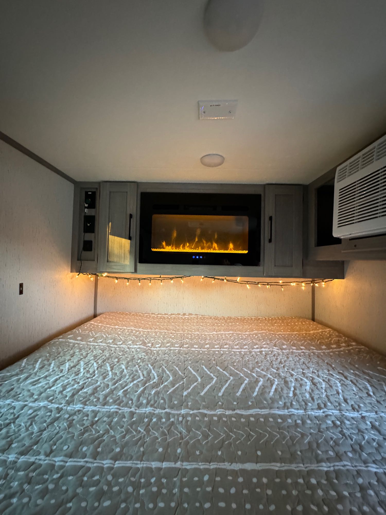 Cozy RV rental bedroom interior with a patterned bed, illuminated by string lights and an electric fireplace, plus an AC unit.. Dutchmen Other 2023