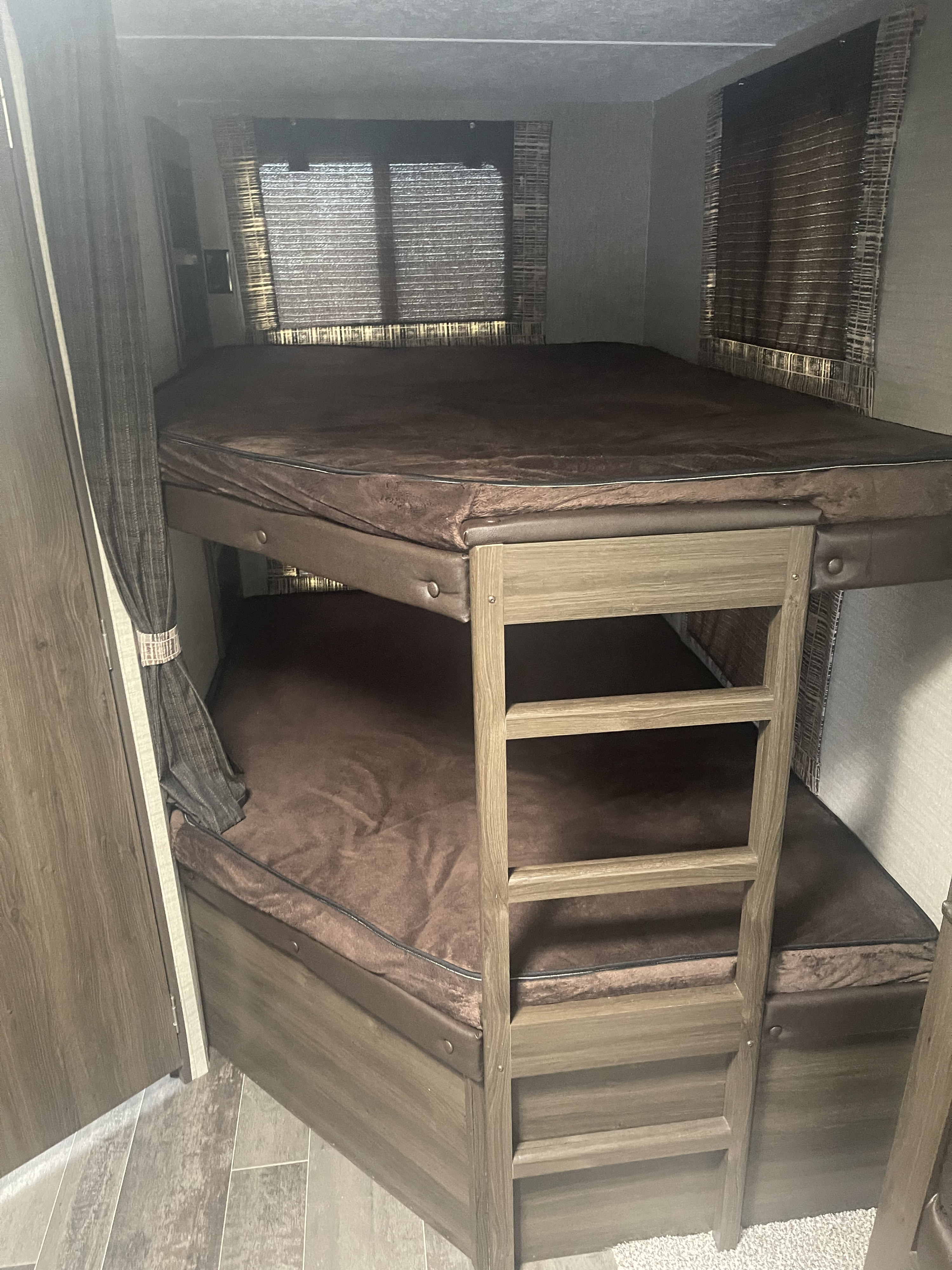 Inside an RV rental, showing built-in bunk beds with dark brown mattresses and a wooden ladder for access. Windows have blinds.. Keystone RV Bullet Ultra Lite 2018