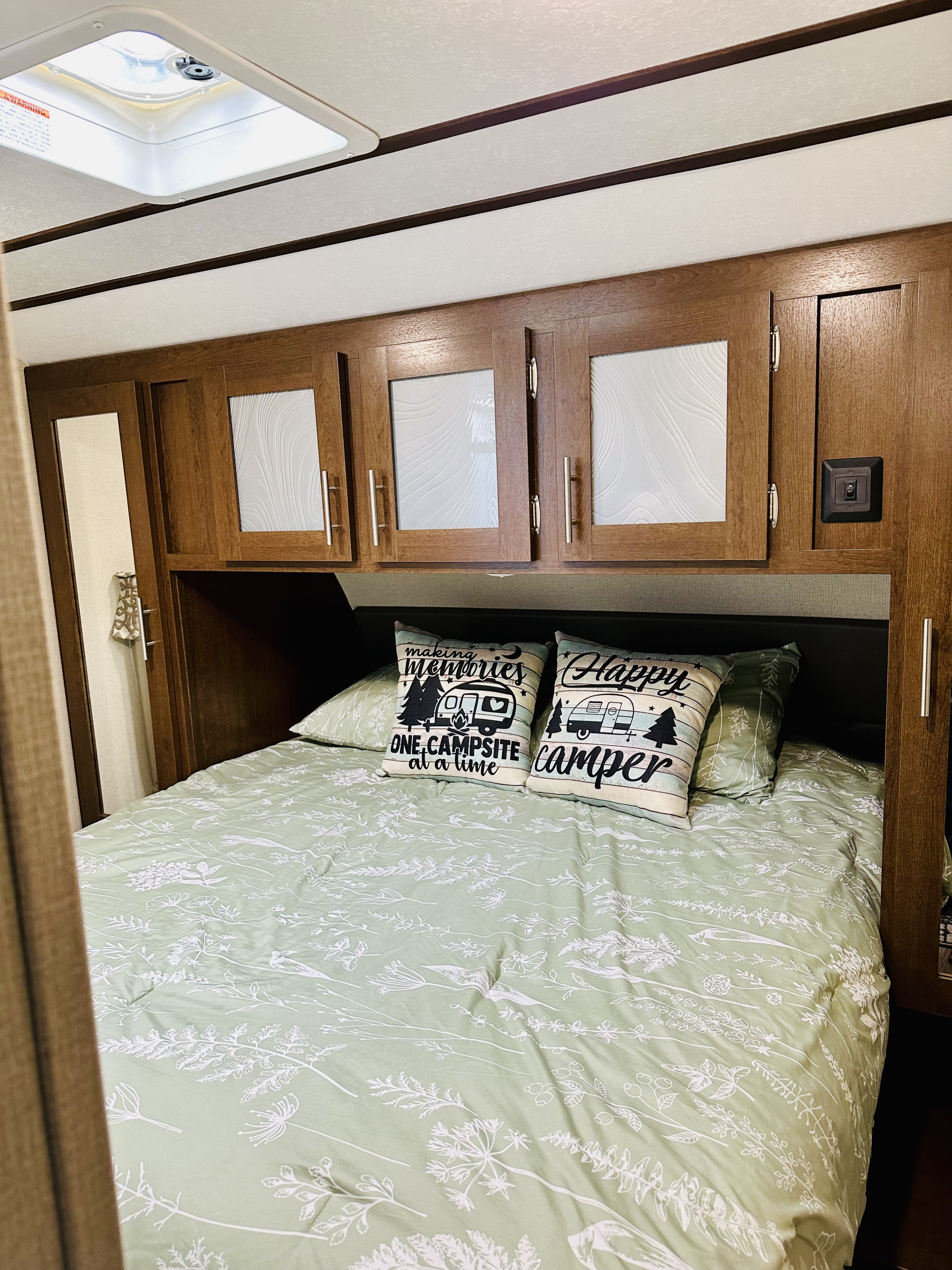 Cozy RV rental bedroom with sage green floral bedding, "Happy Camper" pillows, wood cabinets, and ceiling vent.. Forest River Wildcat 2019