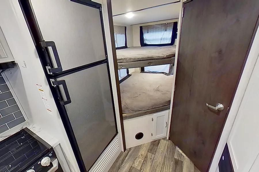 View inside an RV rental showing a compact kitchen with a refrigerator and a sleeping area with bunk beds.. Keystone Bullet 2021