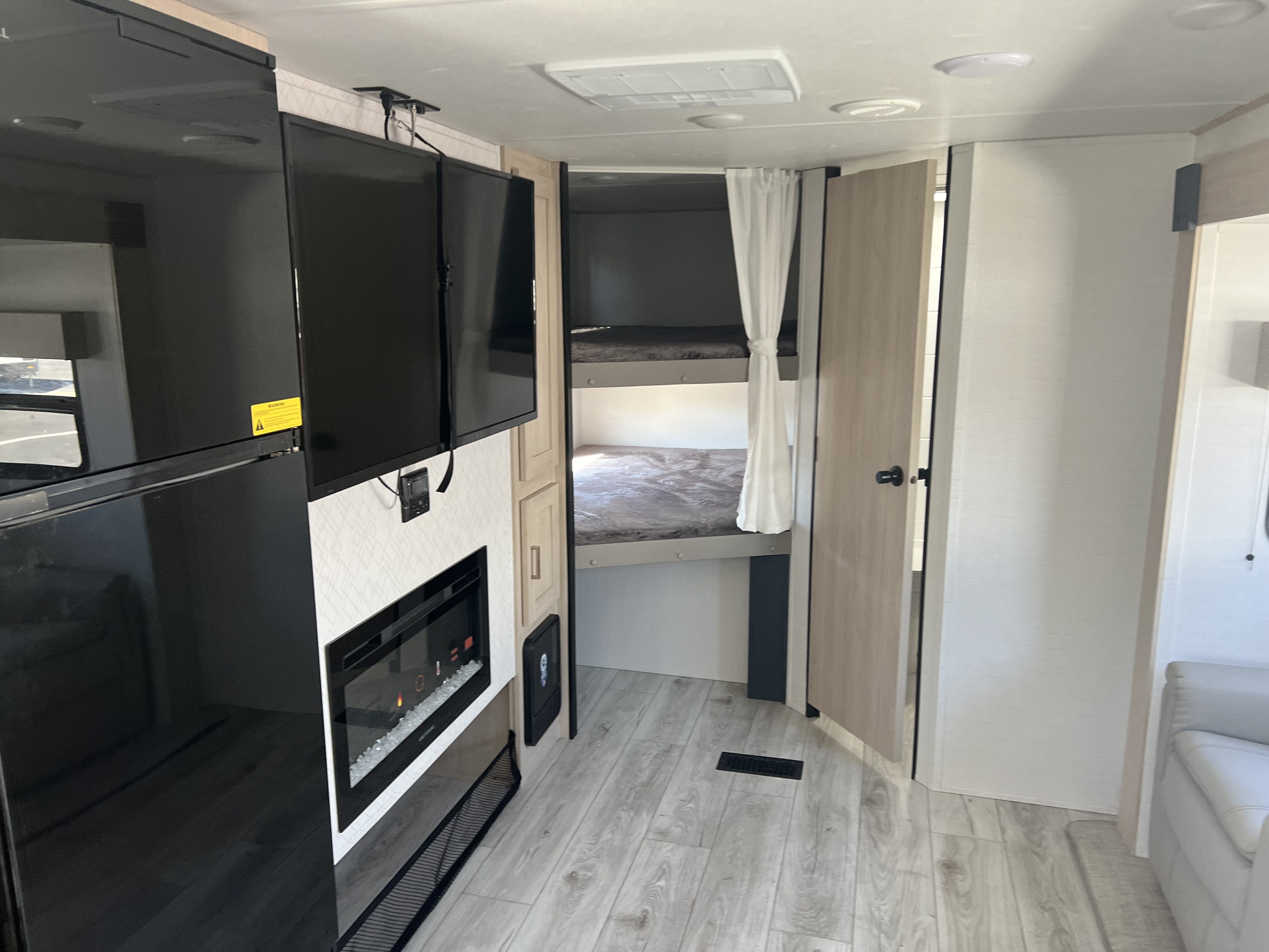 RV rental interior showcasing a large black refrigerator, two TVs, an electric fireplace, and cozy bunk beds with a curtain.. Forest River avenger 2024