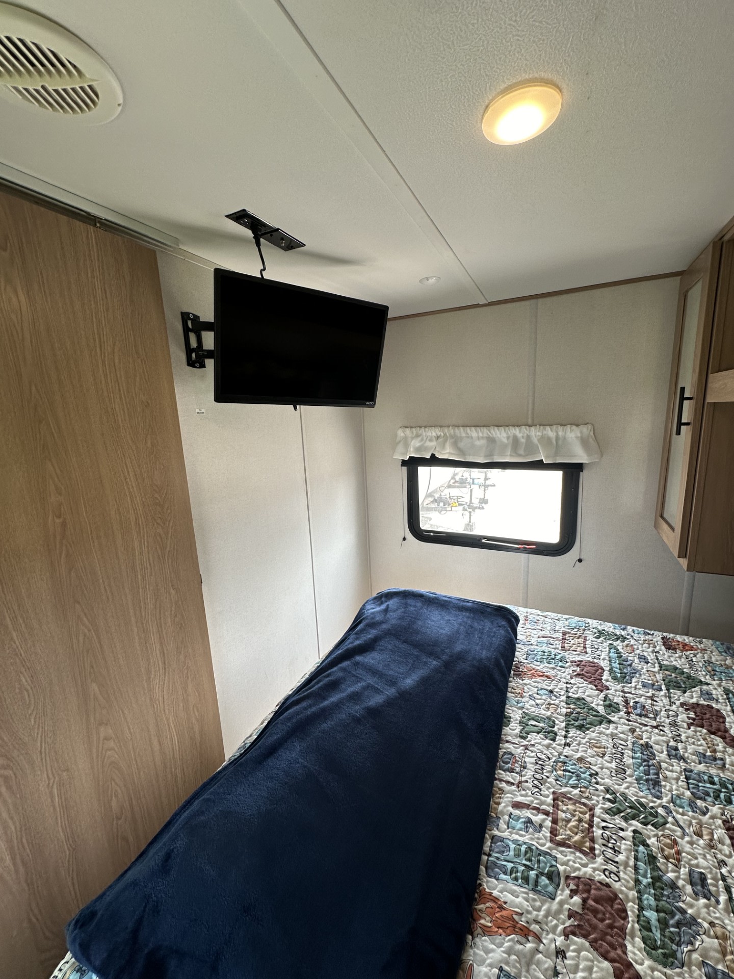 Compact RV rental bedroom with wall-mounted TV, window, and a bed featuring a nature-themed quilt and blue blanket.. Forest River Prime TIME 2022