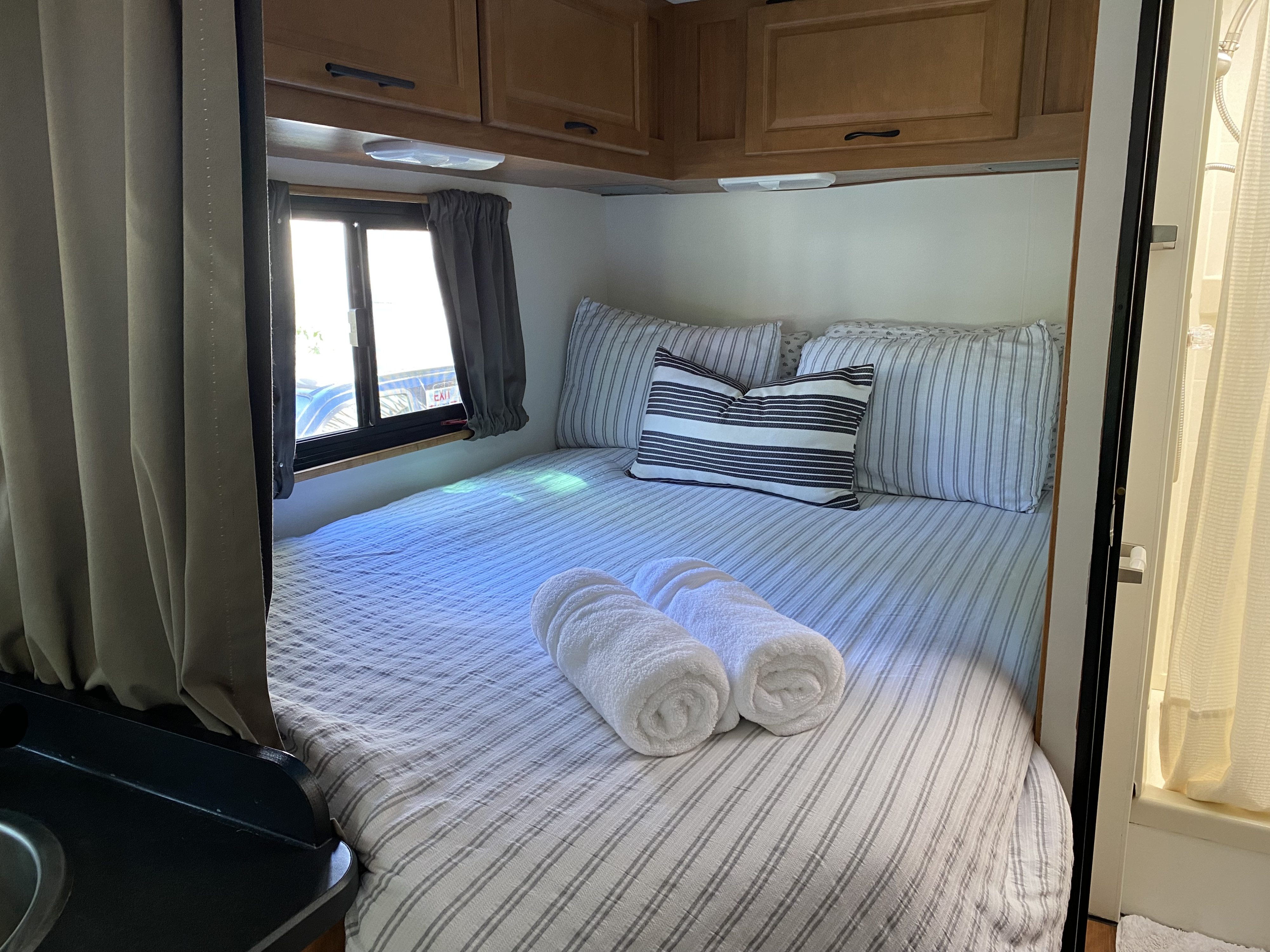 Cozy RV rental bedroom features a comfy bed with striped linens, fresh towels, overhead cabinets, and a window for natural light.. Thor Majestic 2015