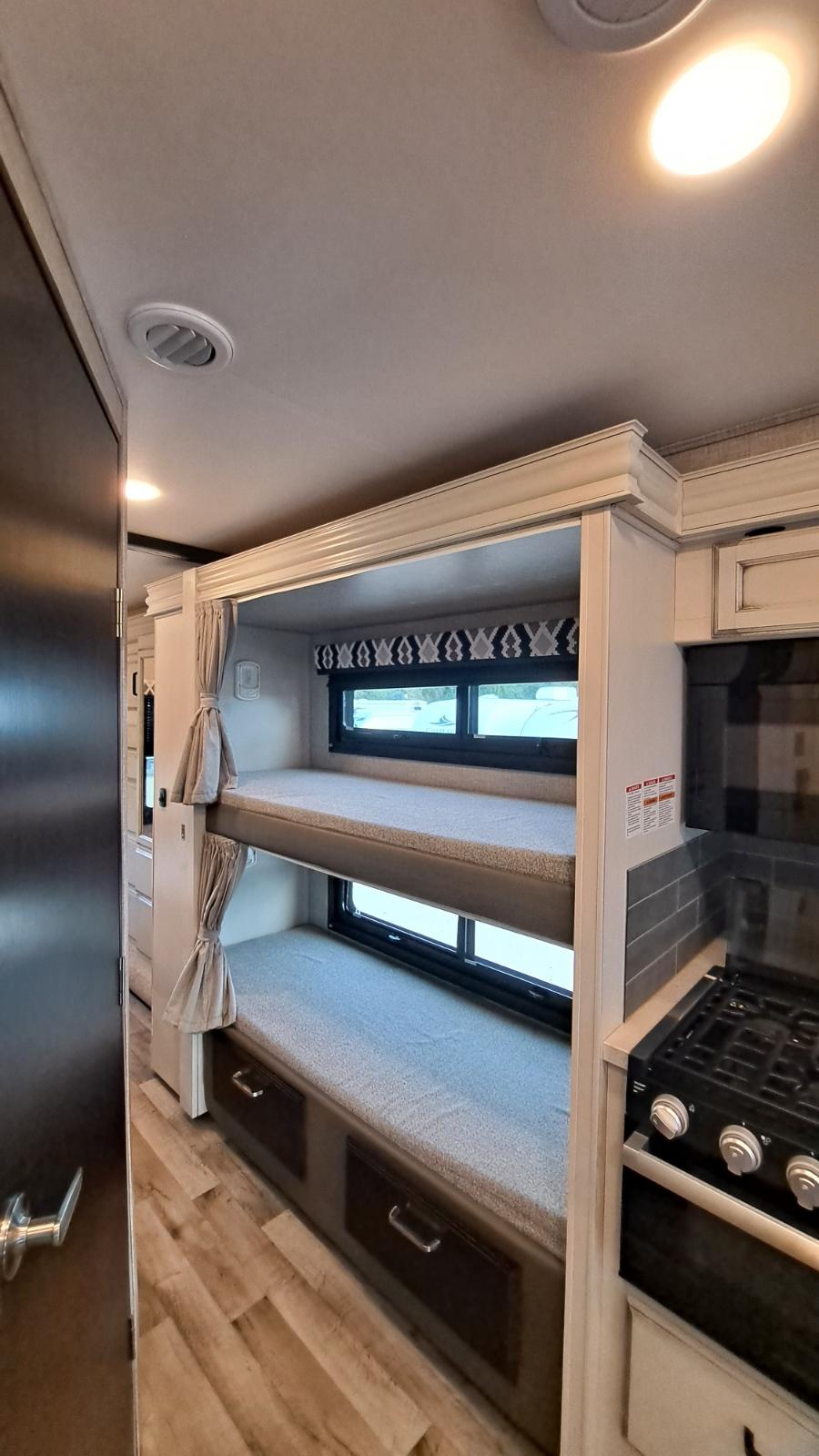 RV rental interior showcasing space-saving bunk beds with windows and curtains, next to a functional compact kitchen.. Jayco 31F 2022