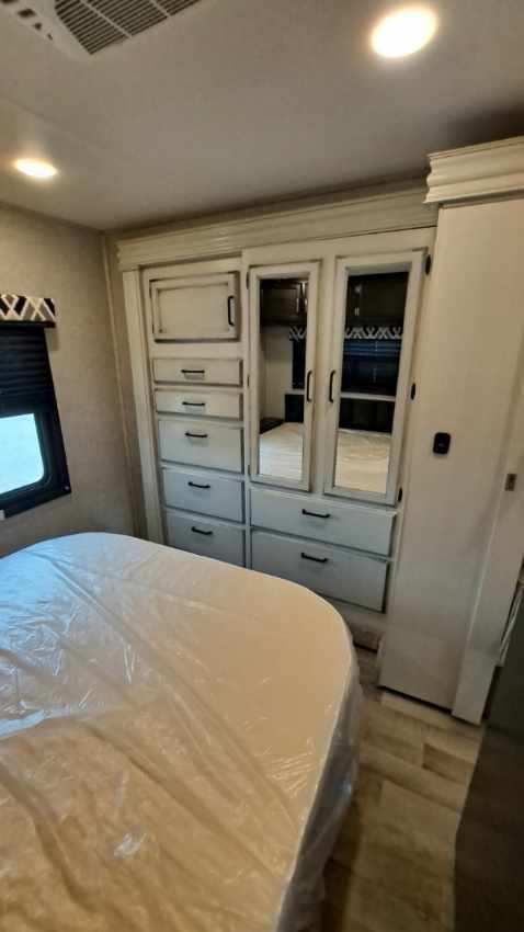 RV rental bedroom: white built-in wardrobe, dresser, mirrored doors, and new bed with plastic cover. Compact interior.. Jayco 31F 2022