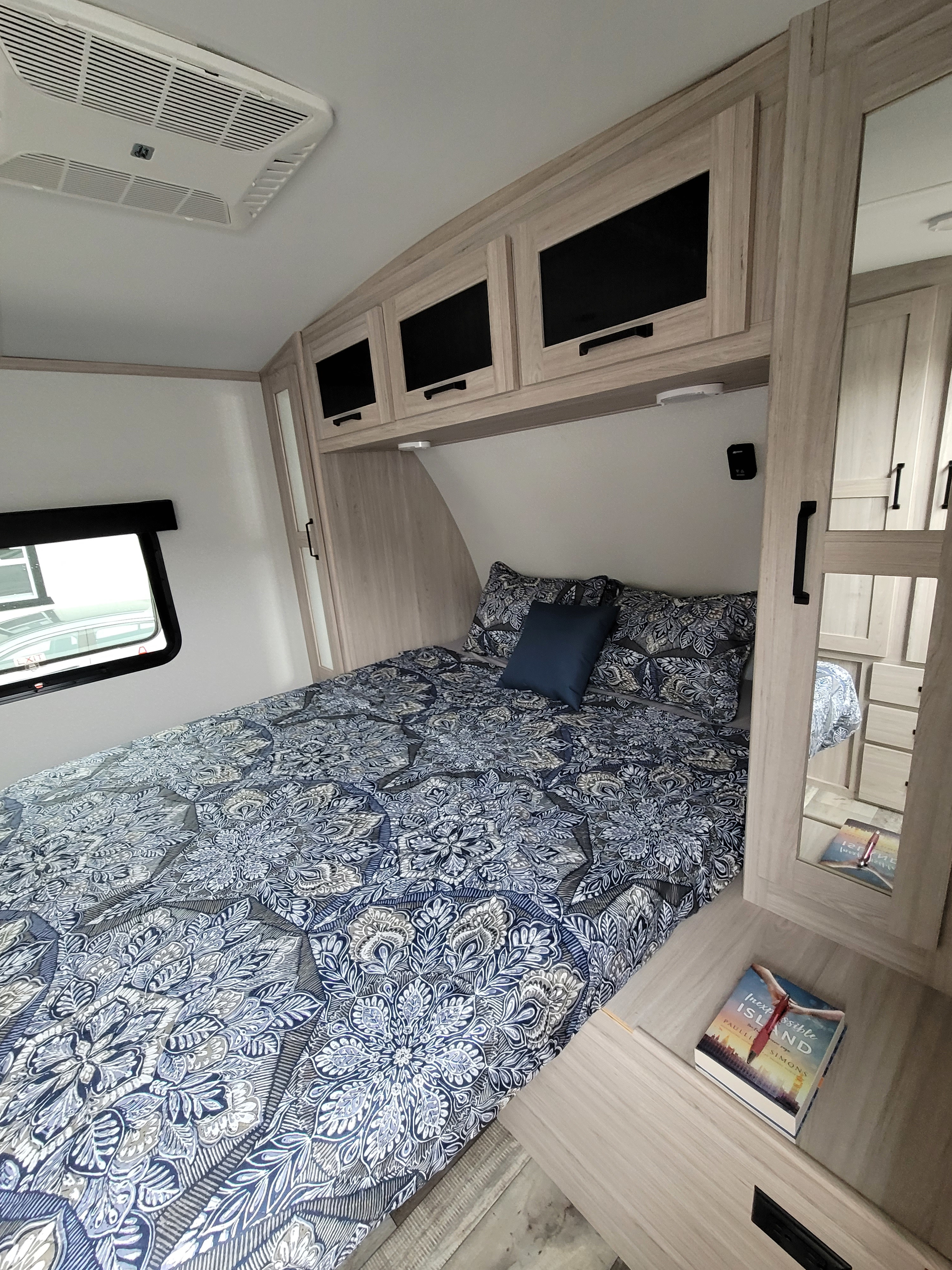 Inside an RV rental, this cozy bedroom features patterned blue bedding, overhead cabinets, and a ceiling AC unit.. Heartland RVs Sundance Ultra Lite 2022
