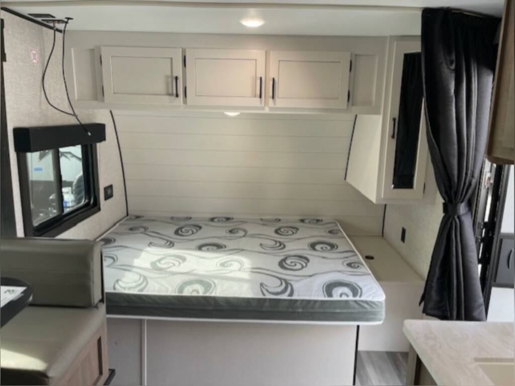 View of a clean RV rental bedroom area with a patterned mattress, white shiplap walls, overhead cabinets, and an adjacent dinette.. Starcraft Autumn Ridge 2024
