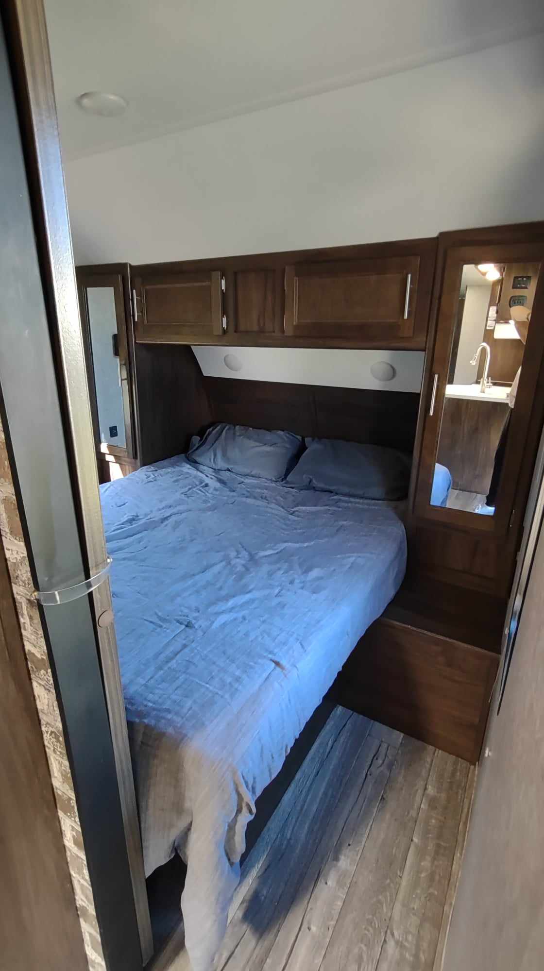 This RV rental shows a compact bedroom with a bed, blue bedding, overhead cabinets, and mirrored storage, reflecting a kitchen.. Forest River Cherokee Grey Wolf 2019
