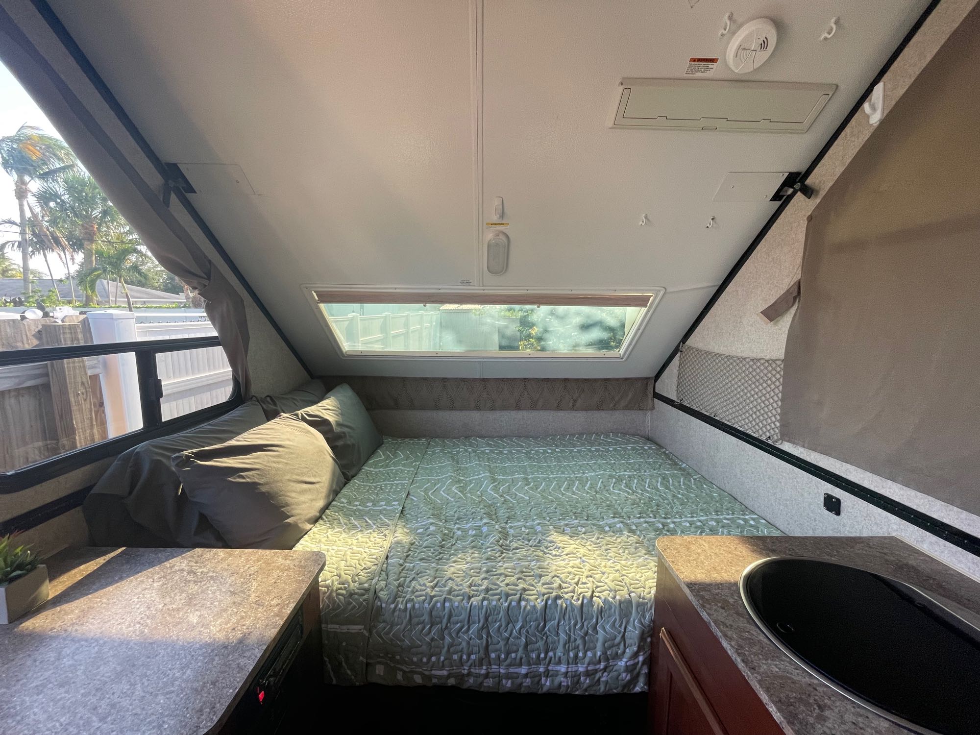 Inside this RV rental: a comfy bed with green quilt, counter space, sink, and sunny windows. Great for camper life!. Forest River Flagstaff T19QBHW 2017