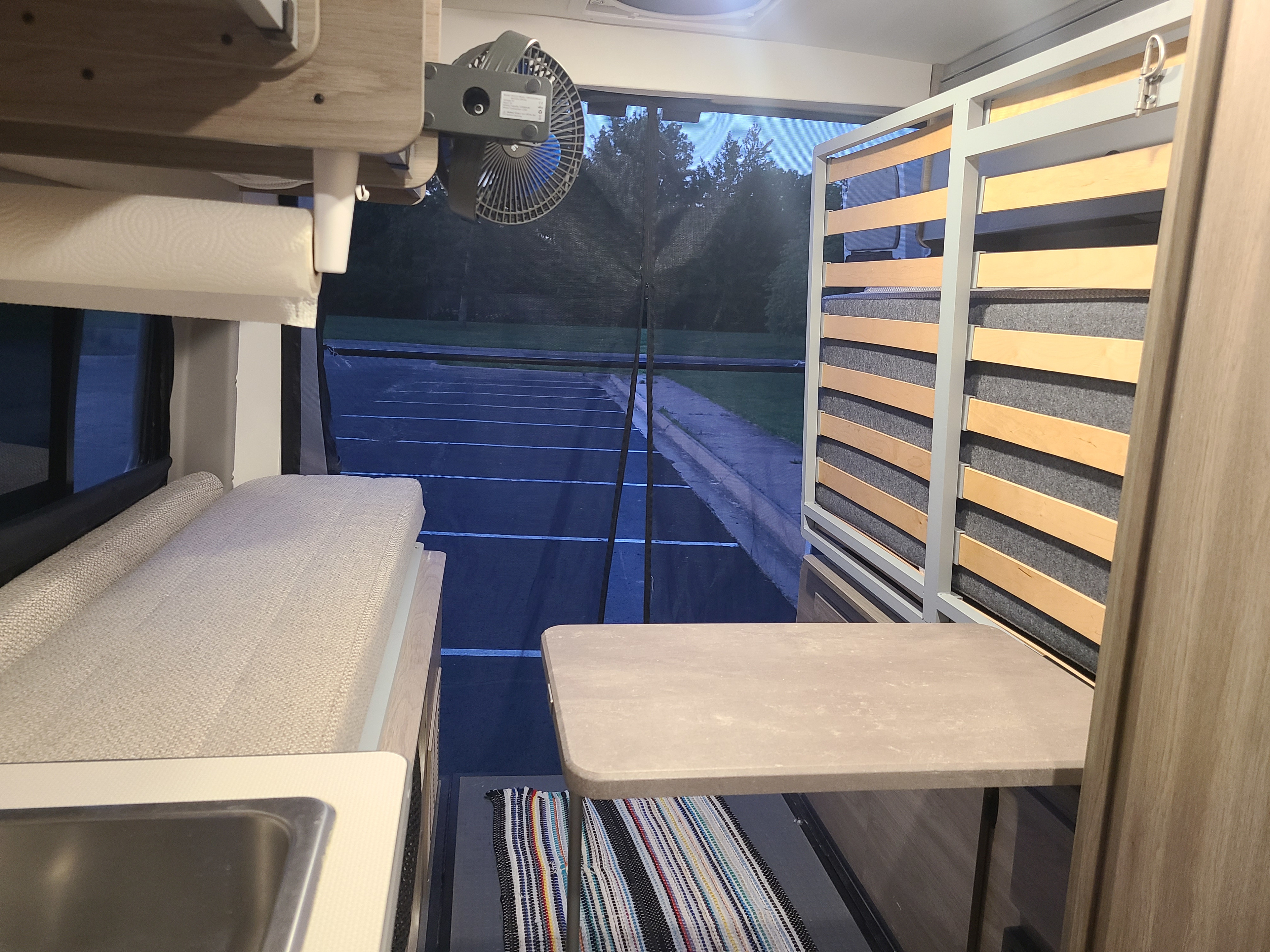RV rental interior featuring a cozy bed, table, fan, and kitchen area, looking out to a parking lot.. Winnebago Solis 2021