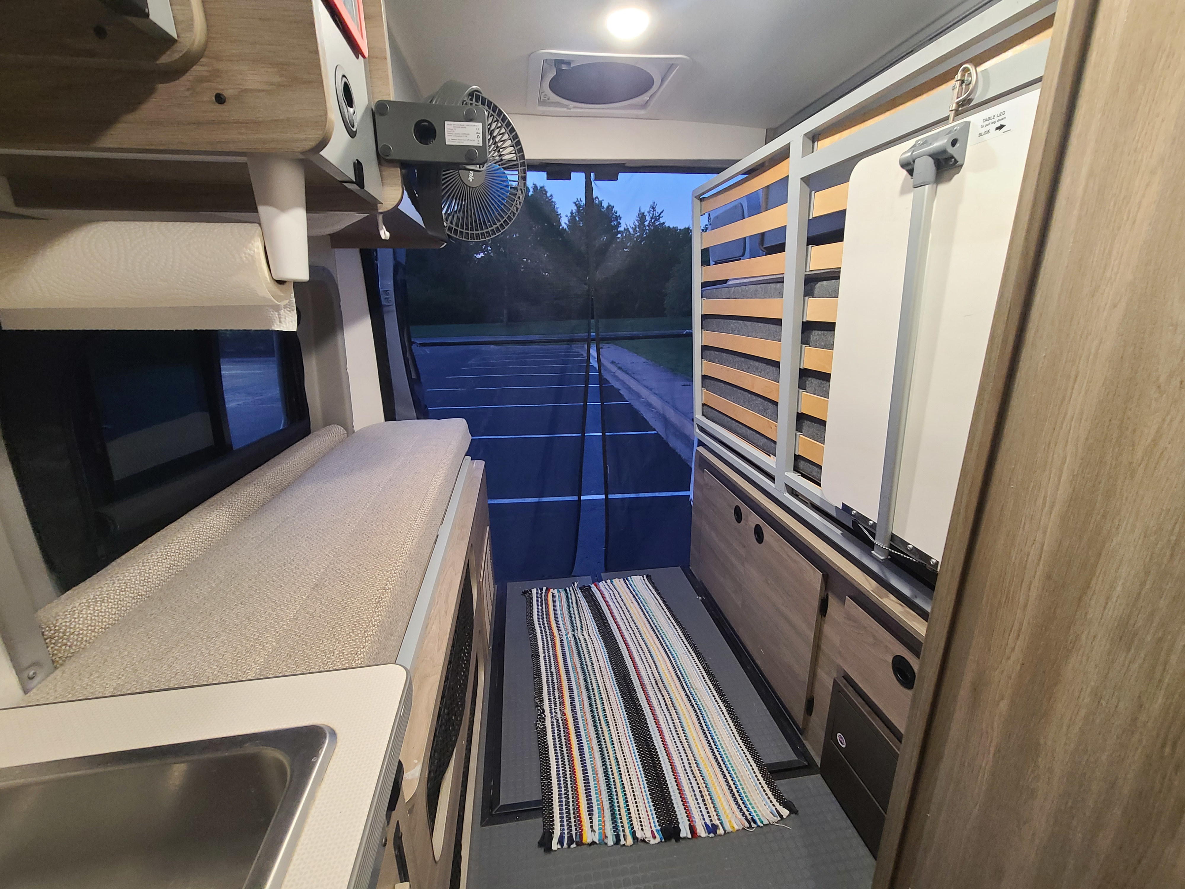 Inside a cozy RV rental: kitchen sink, bench bed, fan, slatted bed frame, & open rear view to a parking lot.. Winnebago Solis 2021