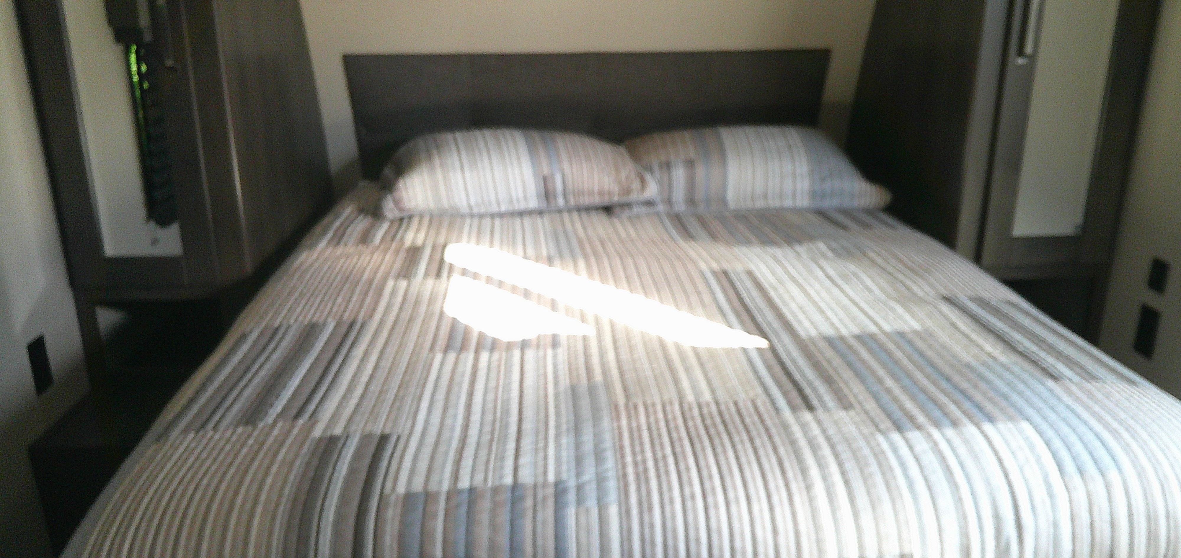 RV rental bed with striped bedding & built-in storage. Bright sunlight on the bed highlights this compact, cozy sleeping area.. Grand Design Explor 2022