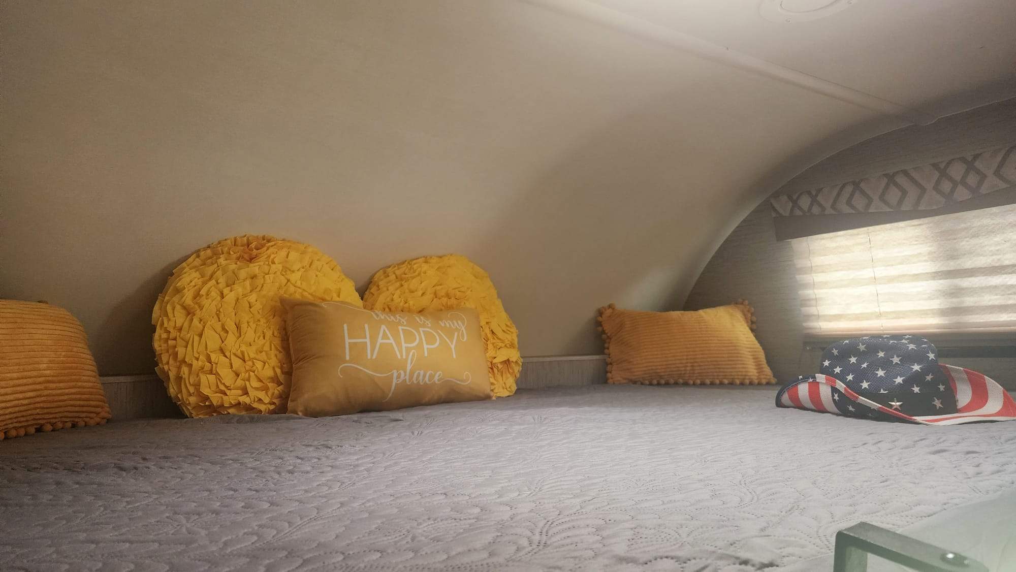 Cozy RV rental bedroom with a comfortable bed, cheerful yellow pillows, one reads 'HAPPY place,' and a patriotic hat.. Thor Quantum 2021