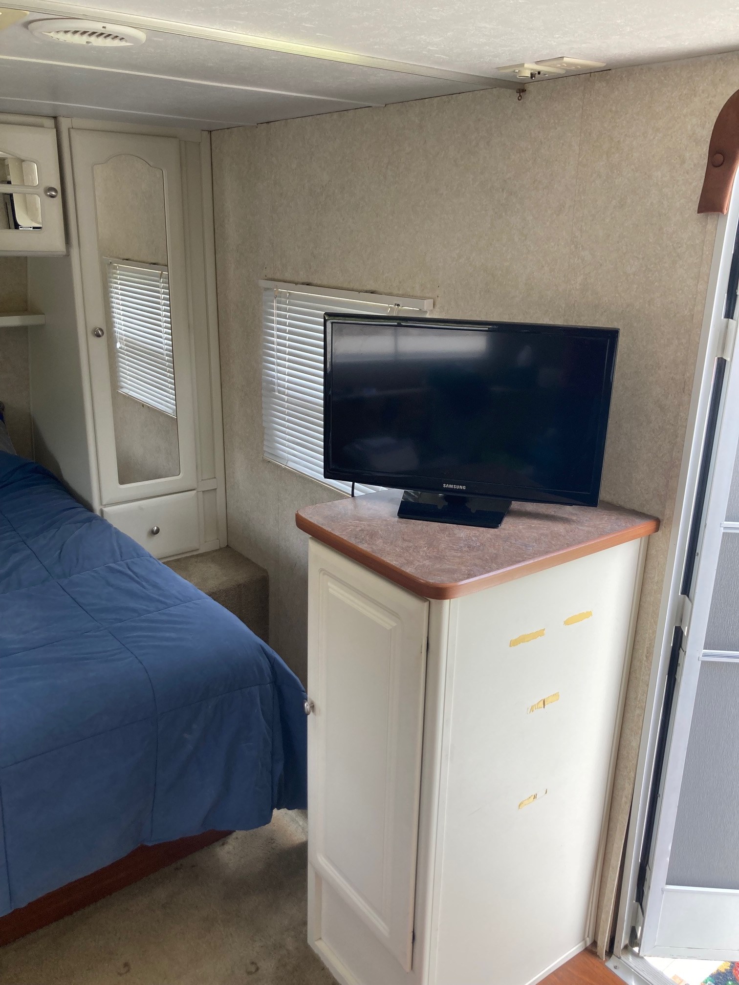 Compact RV rental bedroom featuring a blue bed, TV, and built-in storage cabinet with a mirror.. Keystone RV Outback 2004