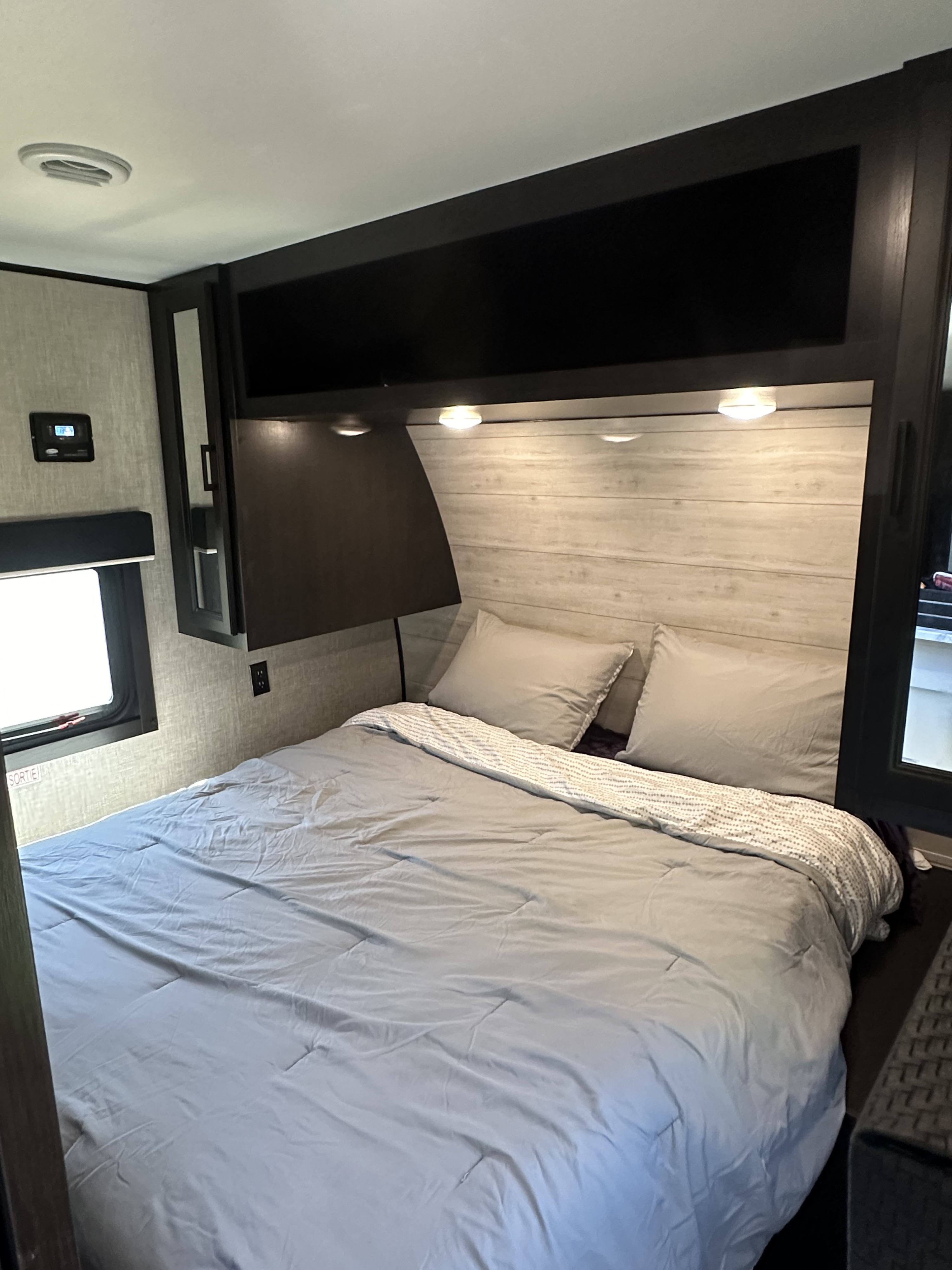 Cozy RV rental bedroom featuring a light wood headboard, grey bedding, overhead cabinets, and a side window, perfect for comfortable travel.. Jayco Jay Flight 2023