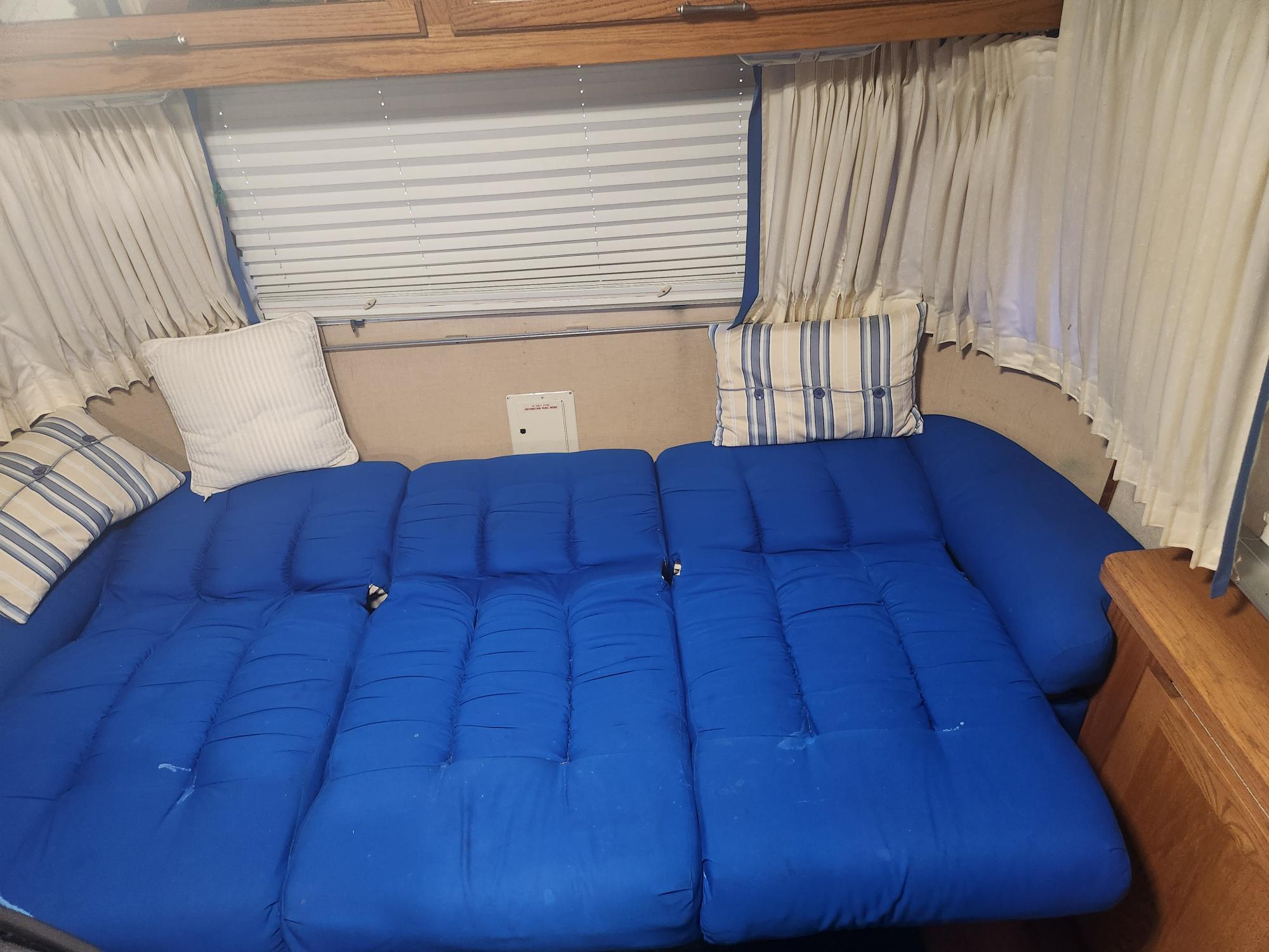 This RV rental interior shows a large bed or seating area with blue cushions, framed by beige curtains, white blinds, and wood trim.. Airstream Limited 2000