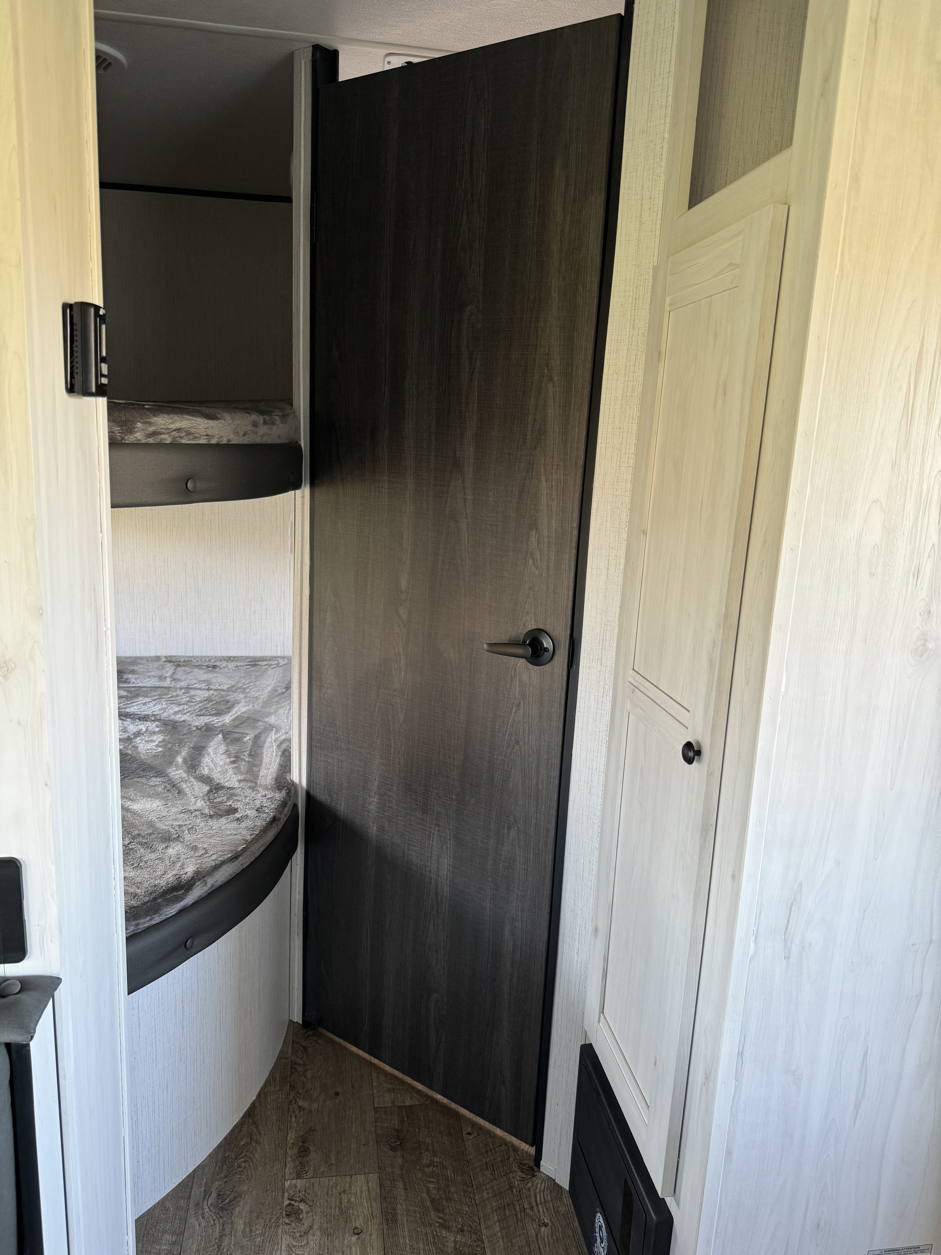Interior of an RV rental: view of a dark wood-grain door, light cabinetry, and two bunk beds with patterned grey covers.. Heartland RVs Trail Runner 2023