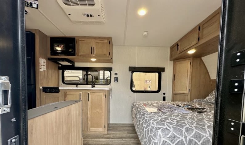 View inside an RV rental: a compact kitchen area with microwave and sink on the left, and a neatly made bed on the right.. Forest River Cherokee Wolf Pup 2023