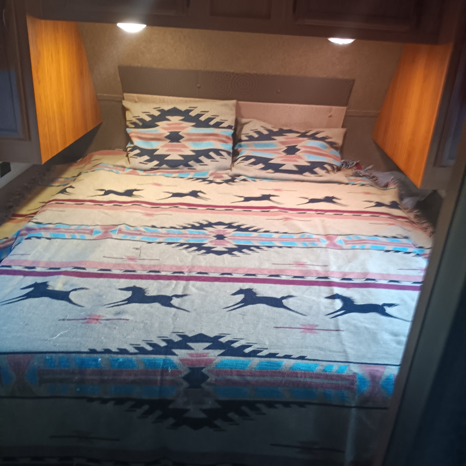 RV rental bedroom: Cozy bed with a vibrant Southwestern-patterned blanket featuring horses and geometric designs. Overhead lights visible.. Starcraft Autumn Ridge 2016