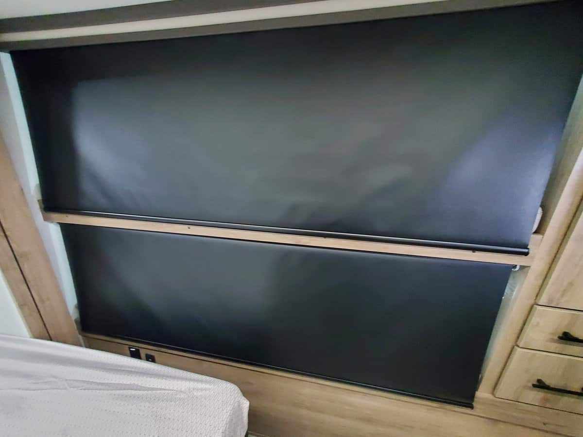 RV rental interior: black roller shades fully pulled down over a window, with bed and wood cabinets nearby.. Grand Design Imagine AIM 2024