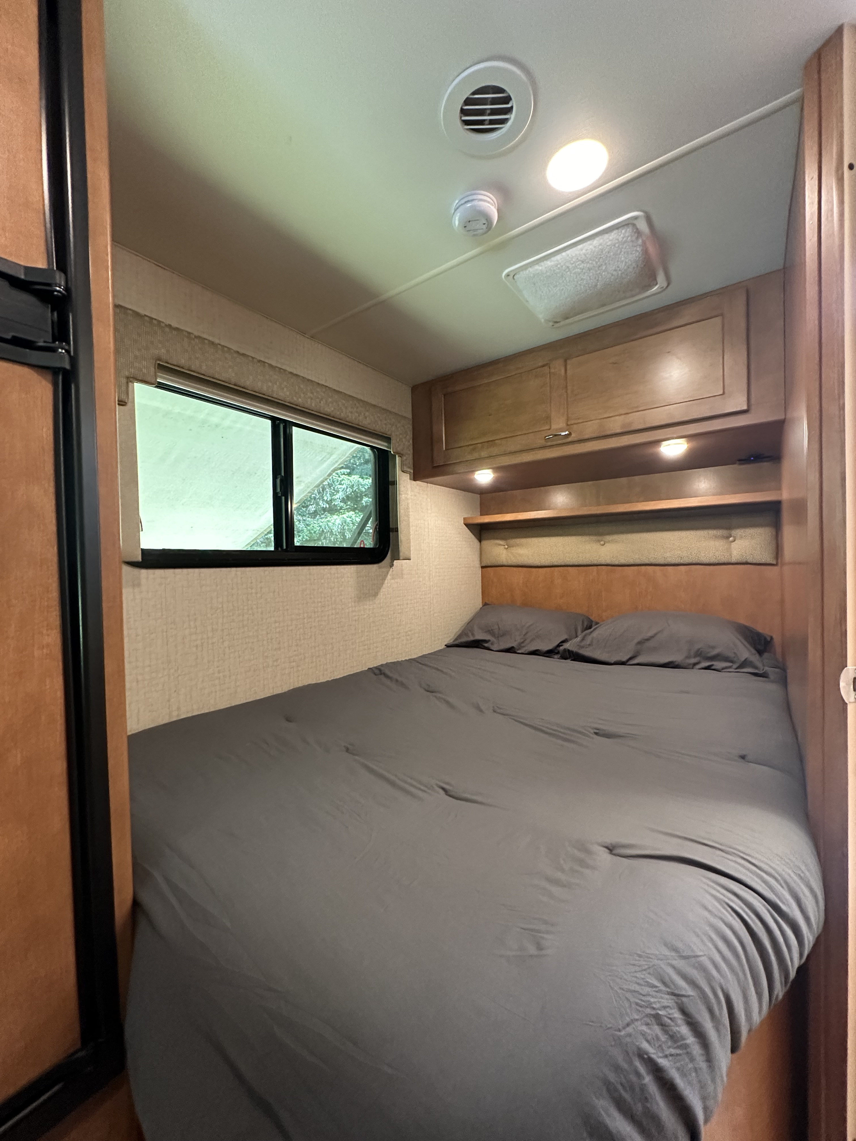 Interior of an RV rental bedroom: bed with gray bedding, wooden overhead cabinets, window, ceiling vent, and lights.. Winnebago Spirit 2017
