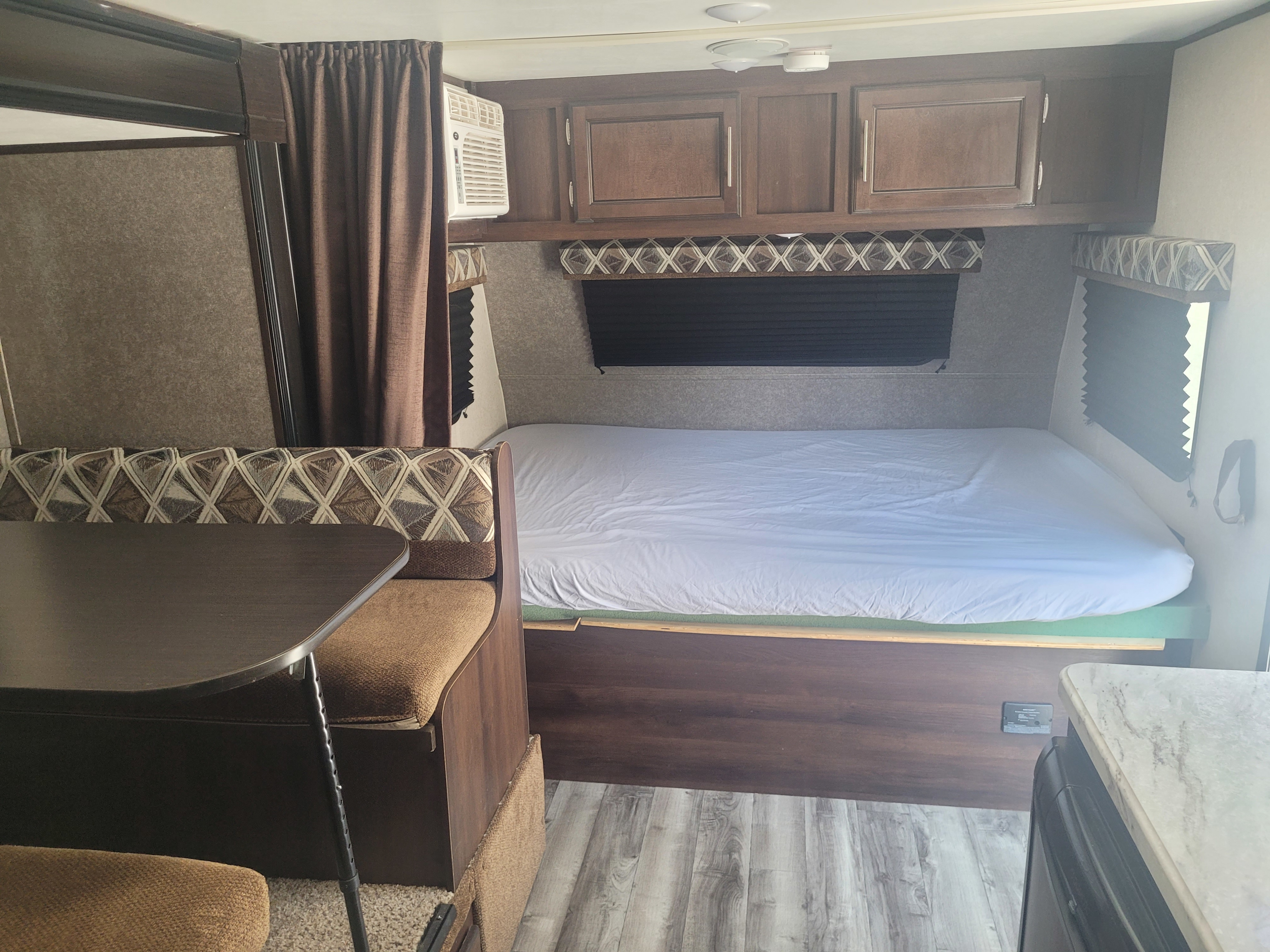RV rental interior: bed with white sheet, dinette seating with patterned cushions, overhead cabinets, and kitchenette.. Jayco Jay Feather 2019