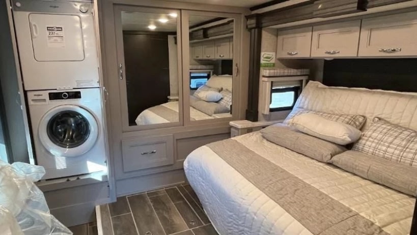 This RV rental bedroom features a stacked washer/dryer, mirrored wardrobe, cozy bed, and overhead cabinets.. Thor Motor Coach aria 2023