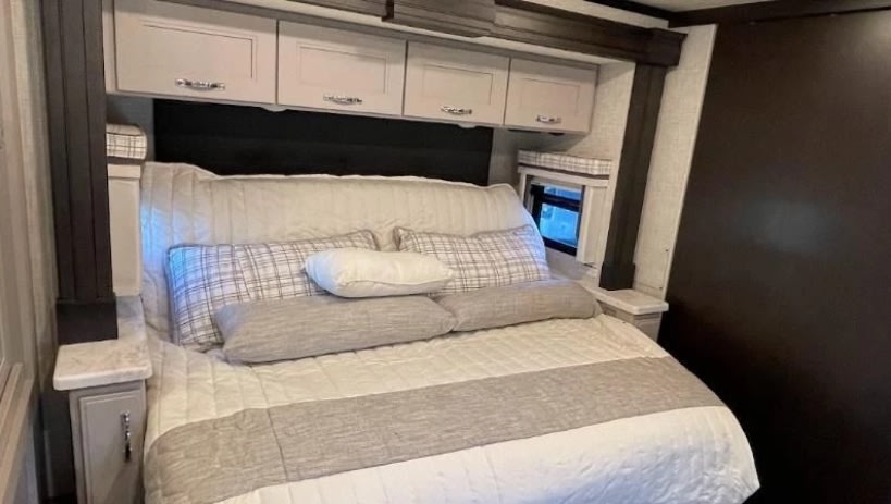 This RV rental bedroom features a comfy queen bed with plaid and gray accents, overhead storage cabinets, and a bedside window.. Thor Motor Coach aria 2023