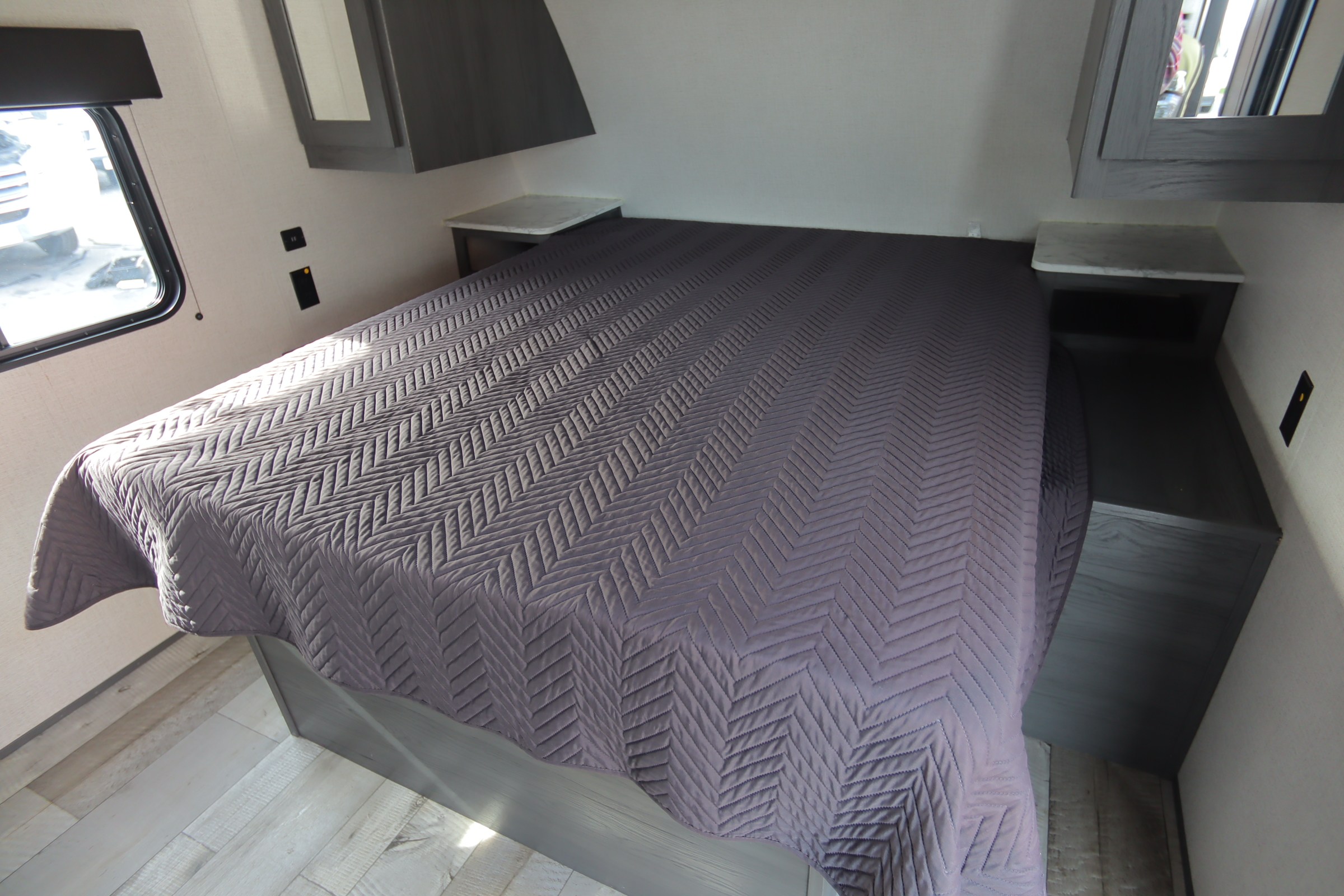 Modern RV rental bedroom: grey quilted bed, dual nightstands, and window, on light wood flooring. Comfortable interior.. Dutchmen Aspen Trail 2022