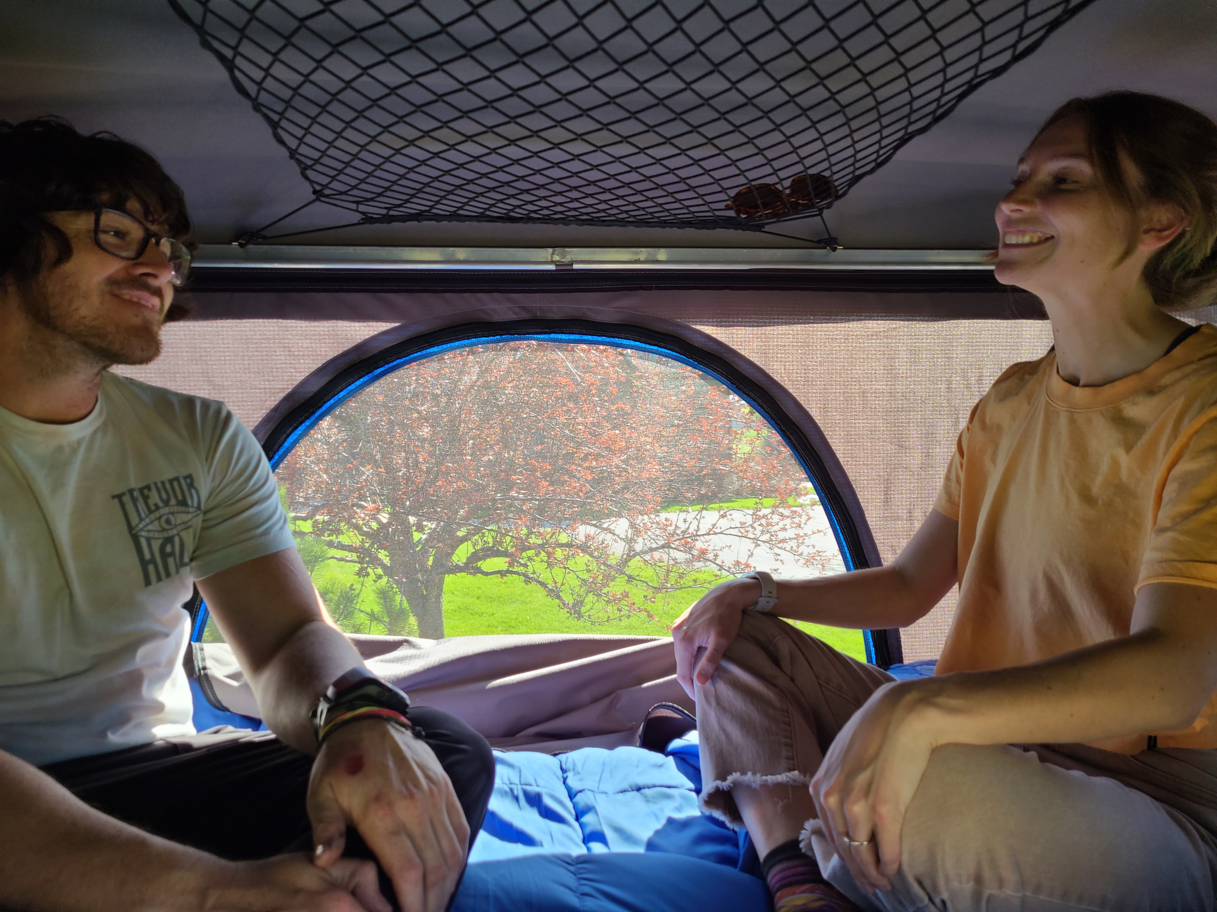 Inside an RV rental, a happy man and woman relax, smiling at each other. Through the window, a tree with reddish leaves and green grass is visible.. Honda Element 2004