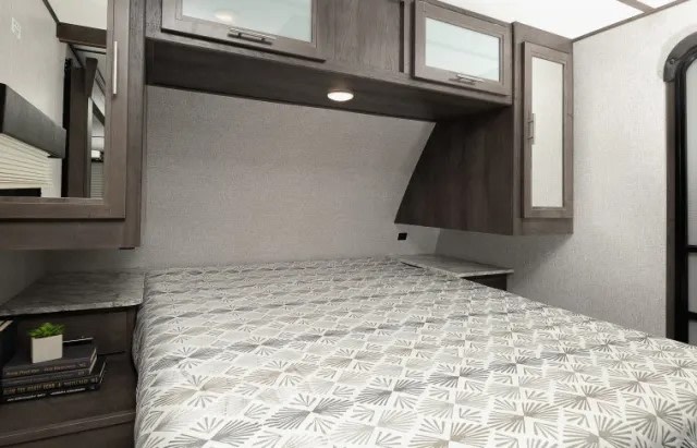 RV rental bedroom: bed with patterned comforter, overhead storage cabinets, and bedside table. Ready for adventure.. Dutchmen Coleman Light 2021