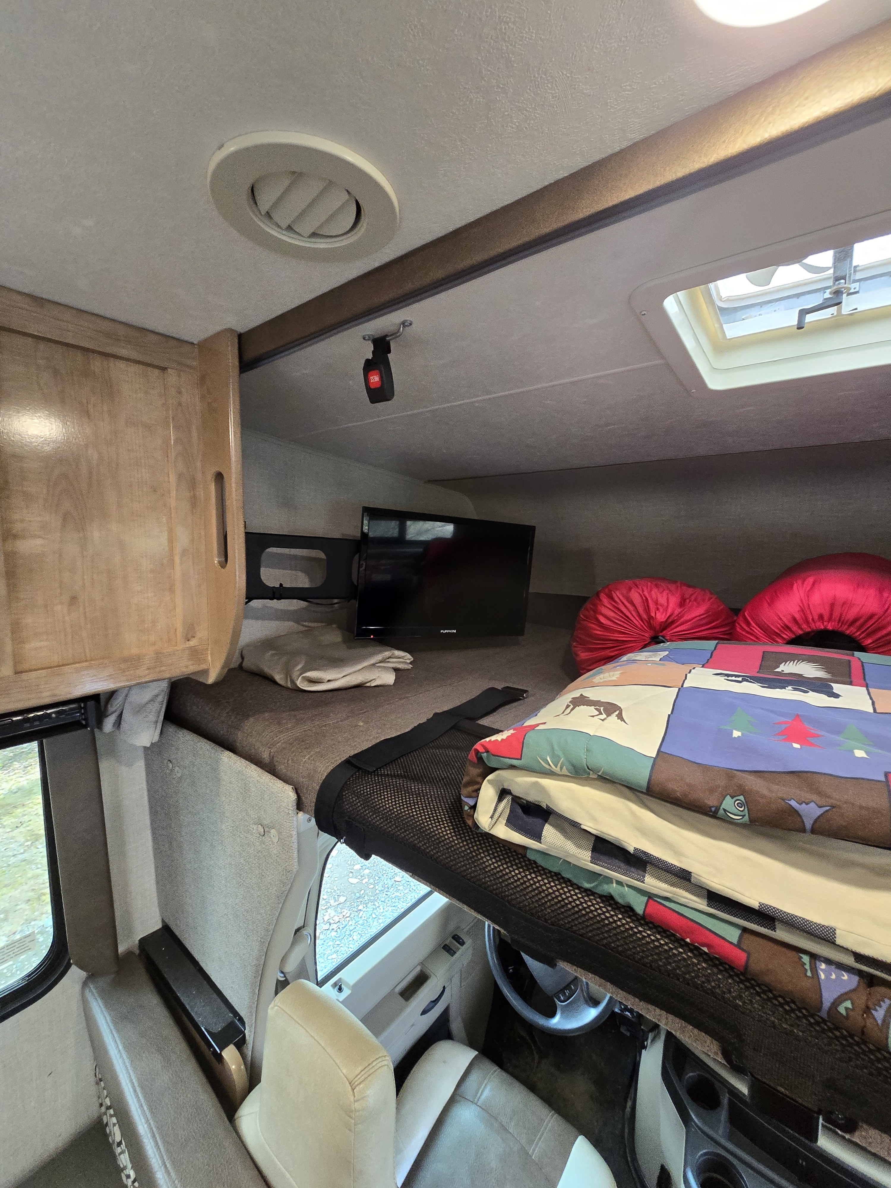 Inside an RV rental: a comfortable cab-over bunk bed with TV, safety net, and skylight. Ready for travel.. Forest River Coachmen Leprechaun 2020