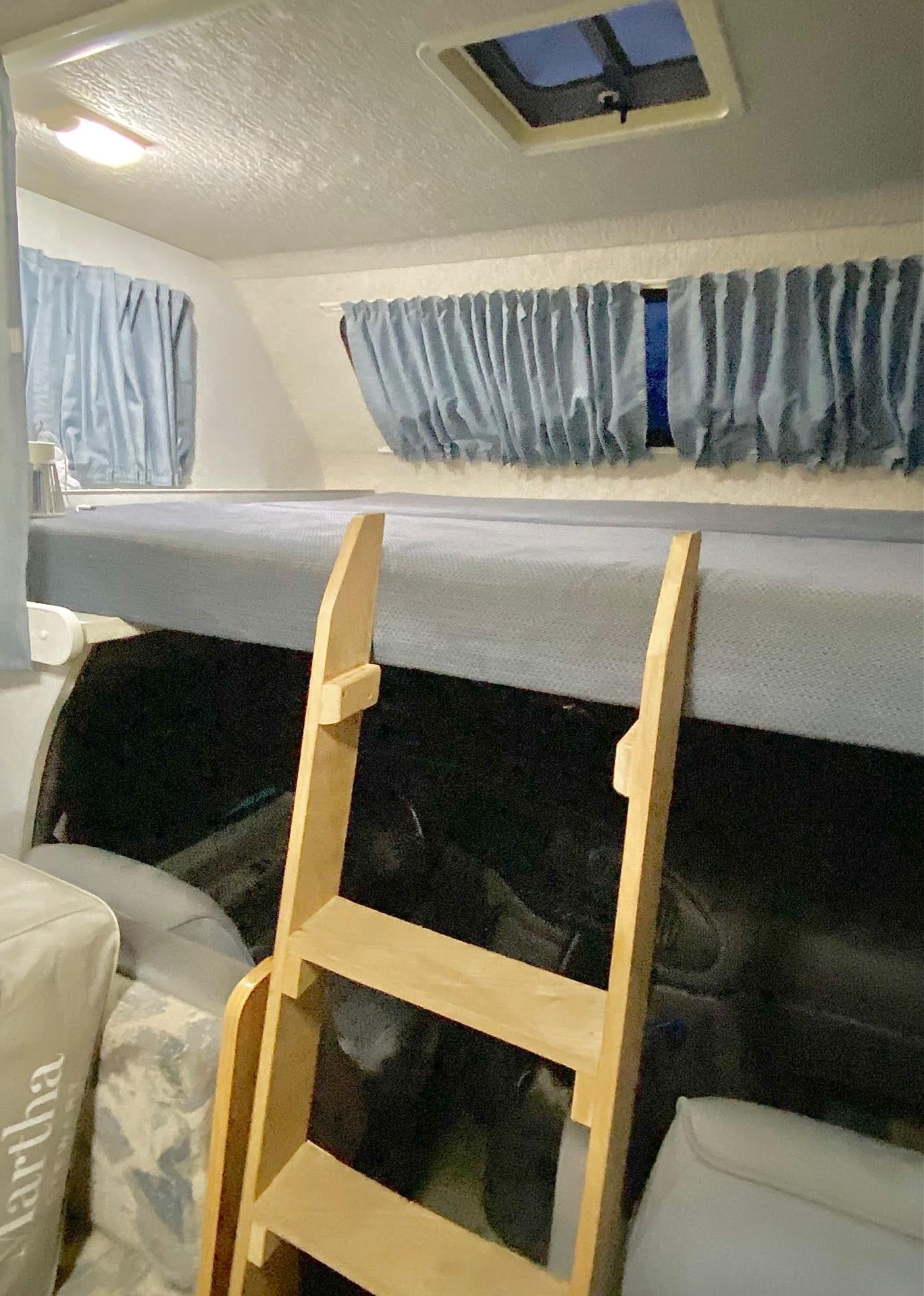 Inside an RV rental, a wooden ladder leads to a bunk bed with a gray mattress, surrounded by blue window curtains and a roof skylight.. Winnebago Minnie Winnie 2001