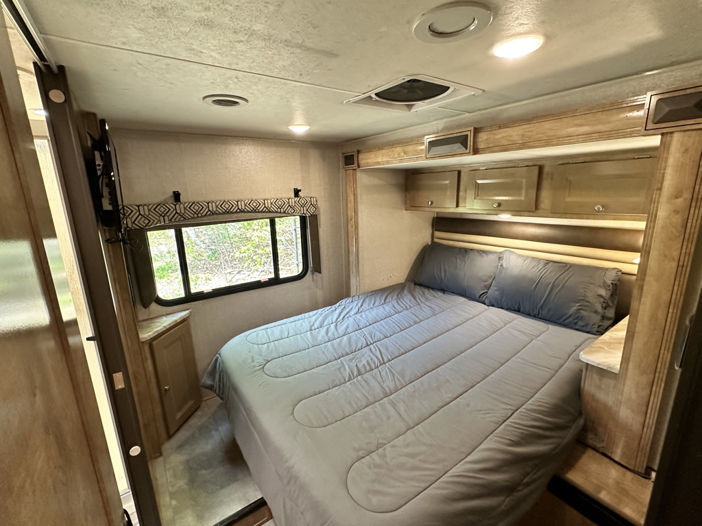 RV rental bedroom interior with a comfy bed, overhead cabinets, window view, and TV.. Coachmen Leprechaun 2019