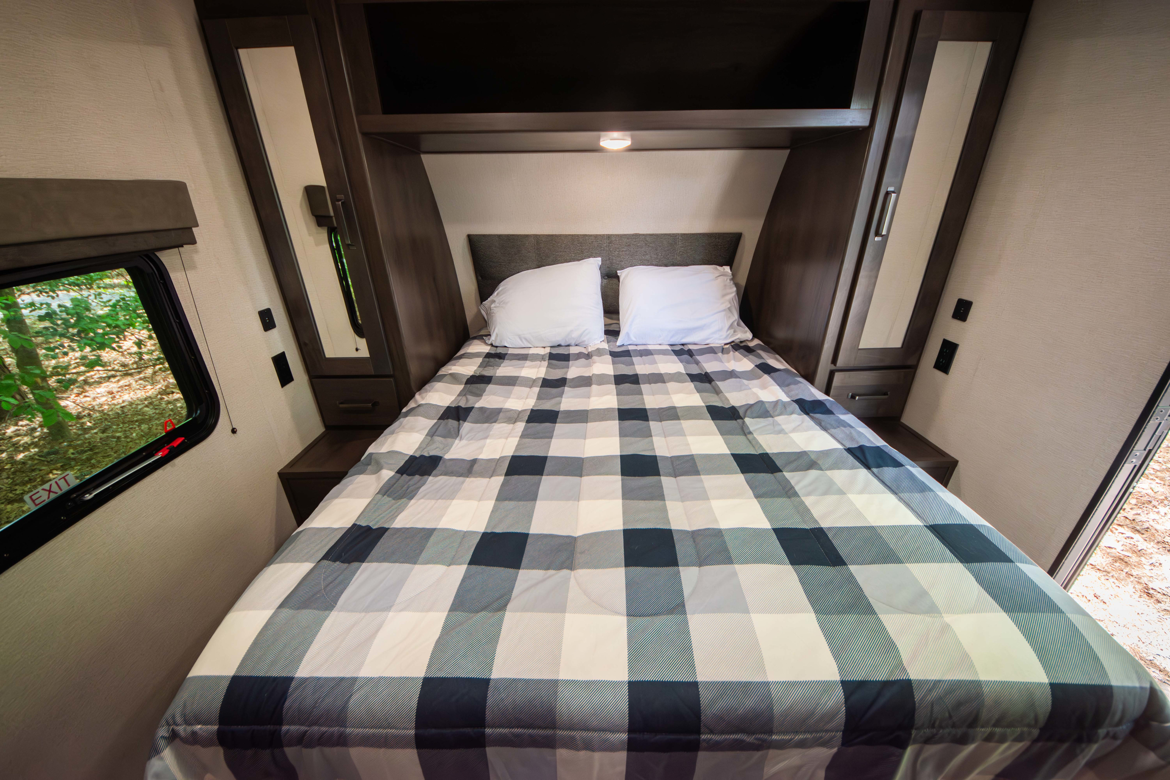 Step inside a comfortable RV rental bedroom, featuring a plaid bed, built-in storage, and a window with an emergency exit.. Grand Design Transcend Xplor 2023