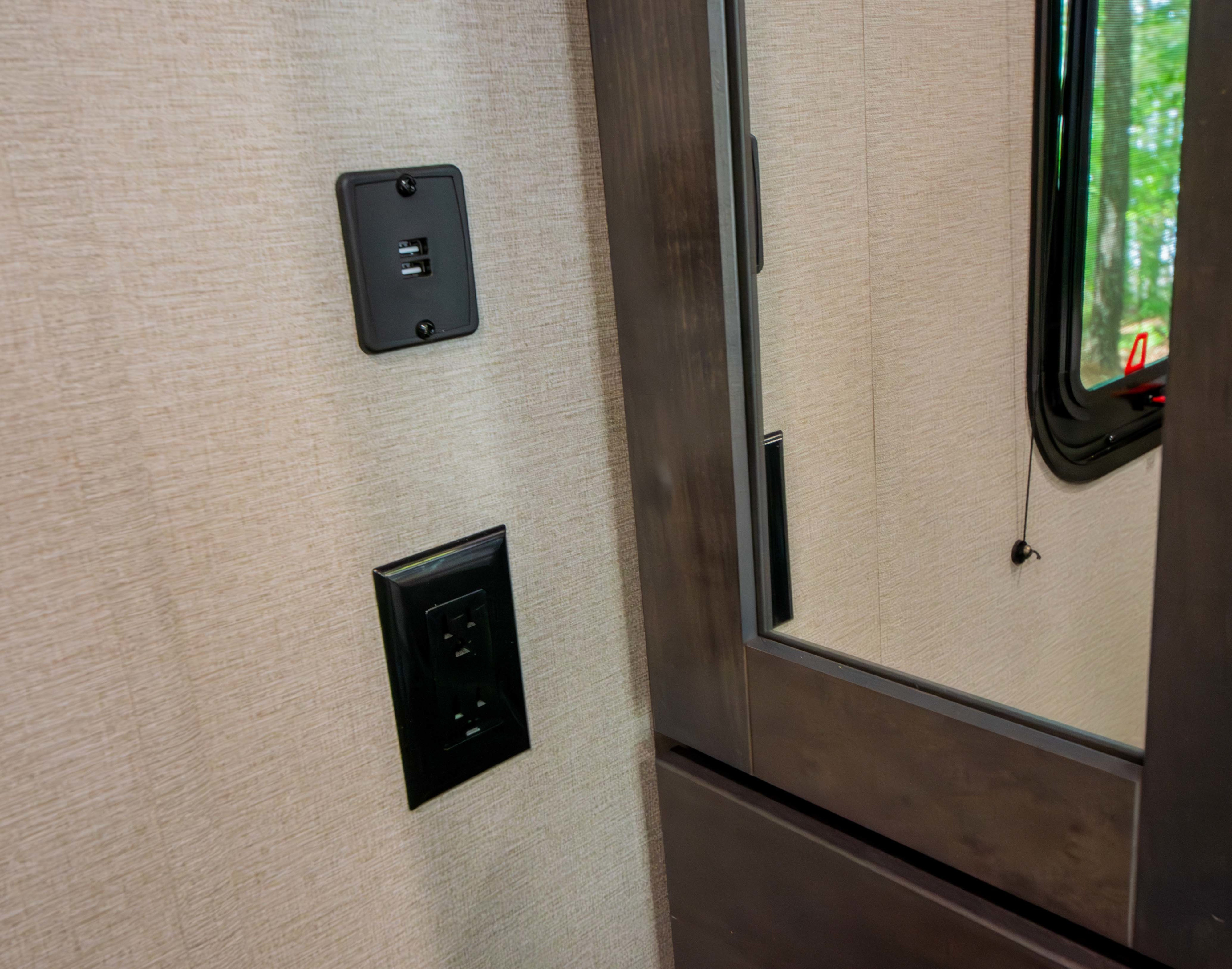 RV rental interior wall: USB charging ports and standard power outlets. Dark wood furniture and window reflection shown.. Grand Design Transcend Xplor 2023