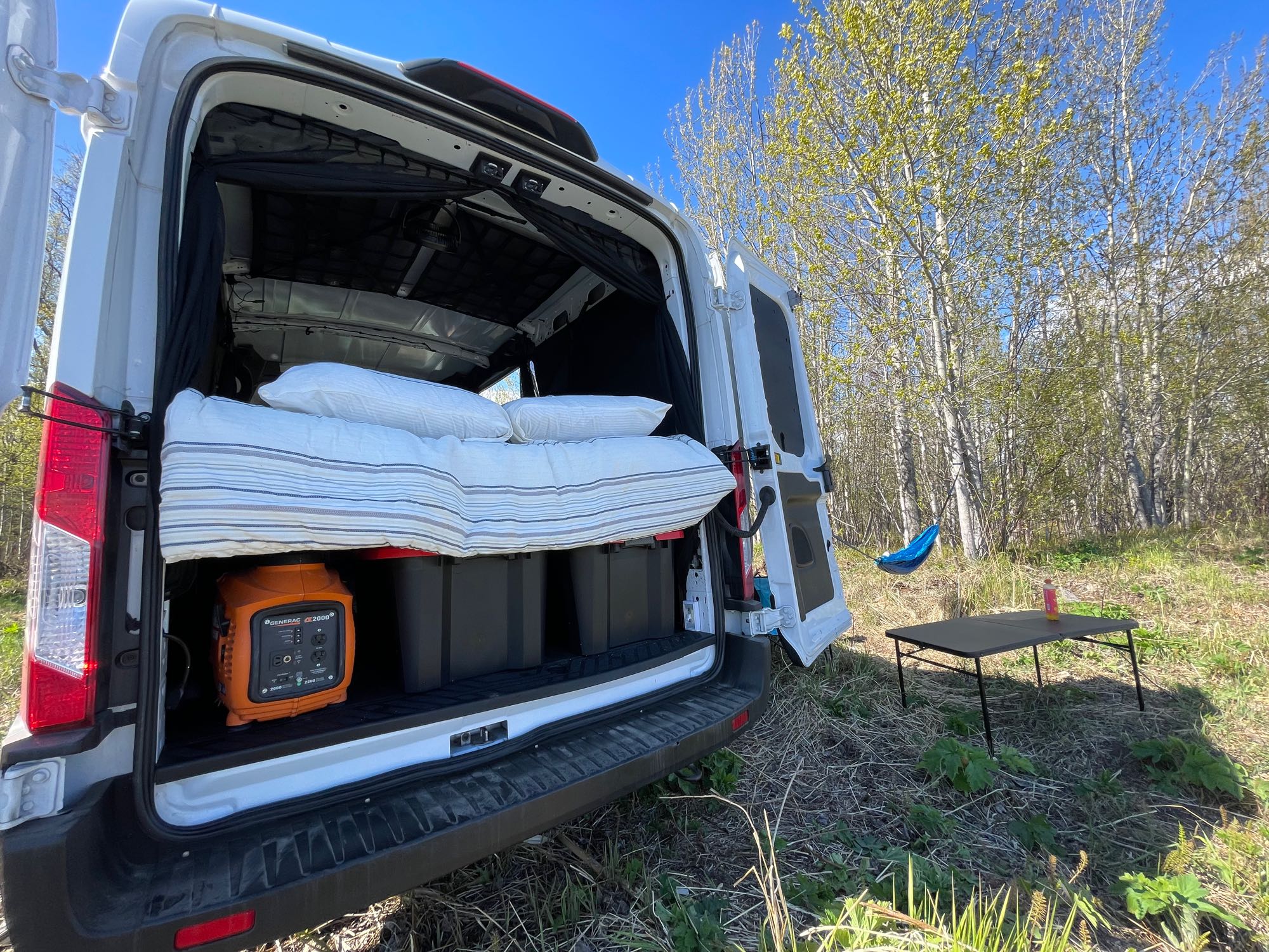 RV rental back doors open to a cozy bed, Generac generator, and storage. An outdoor table and hammock complete this serene camp setup.. Ford Transit 2023