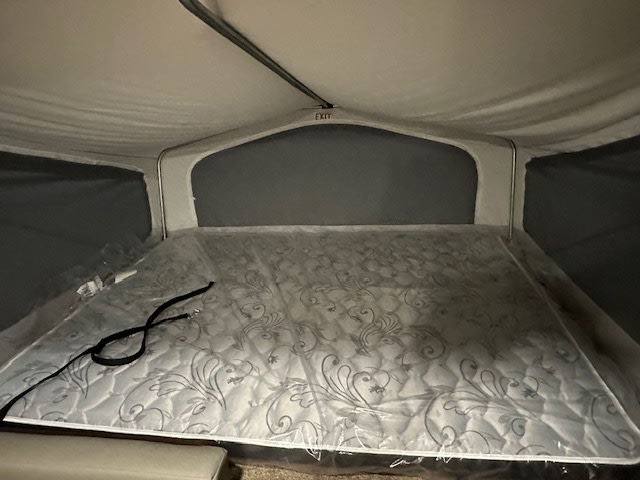 RV rental sleeping area: Pop-up camper bed with patterned mattress under clear plastic. A black strap rests on it.. Solaire 147H 2023