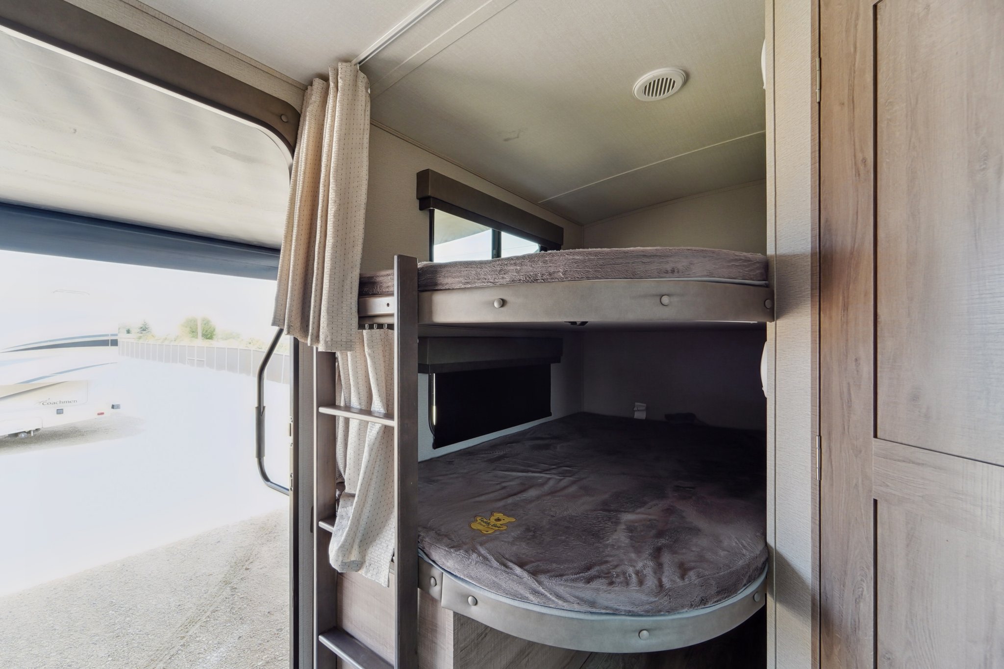 RV rental interior featuring cozy bunk beds with curtains for privacy. A window shows a gravel lot and another RV.. Grand Design Imagine 2021