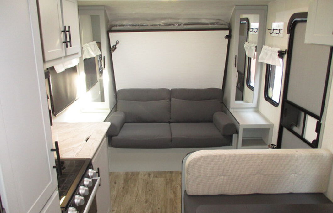 RV rental interior featuring a gray sofa below a fold-down murphy bed, a kitchen counter with stovetop, and cabinets.. Forest River Vibe 2022