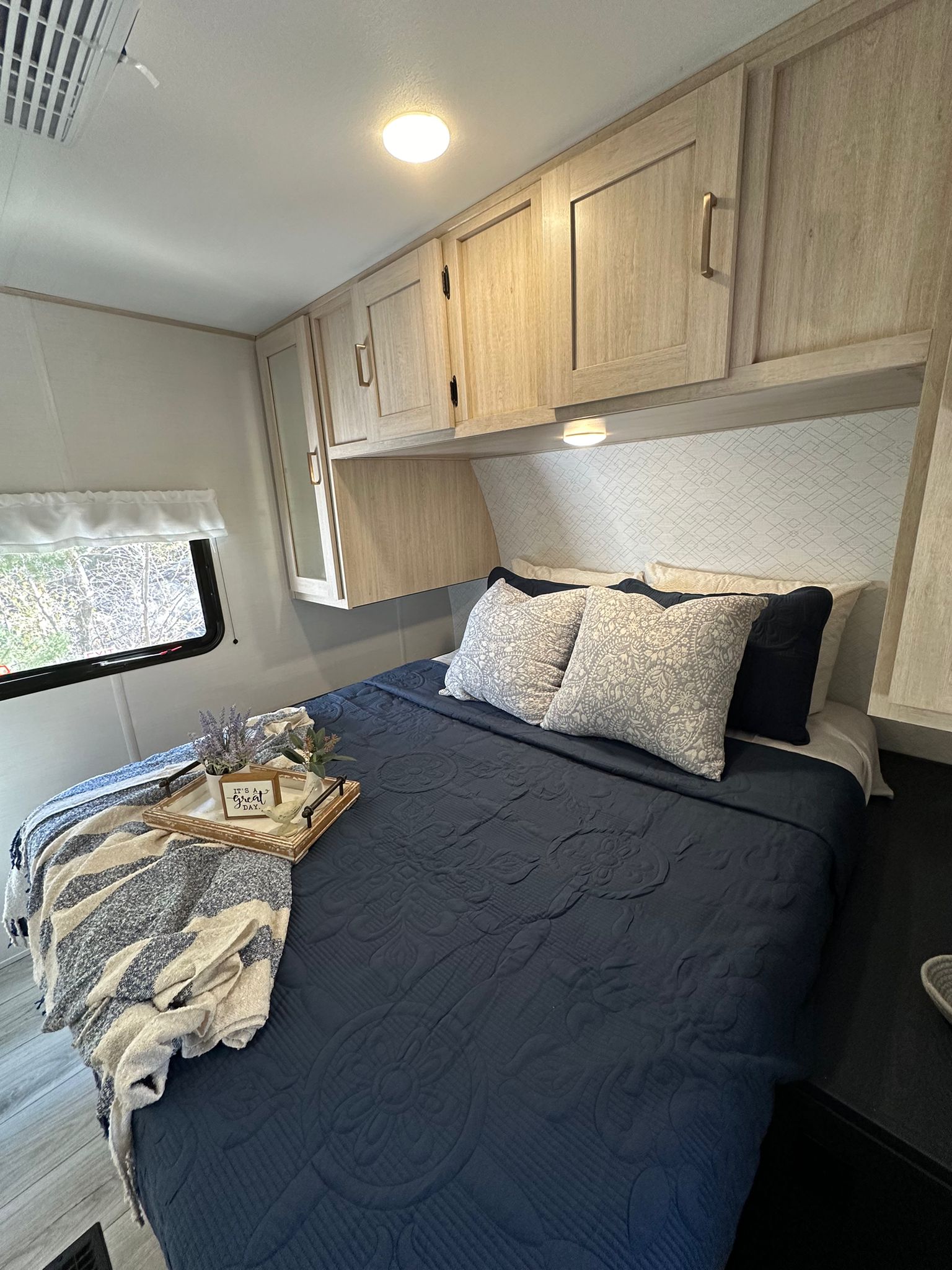 Comfortable RV rental bedroom featuring a bed with navy quilt, decorative pillows, light wood cabinets, and window view.. Avenger ATI 2024
