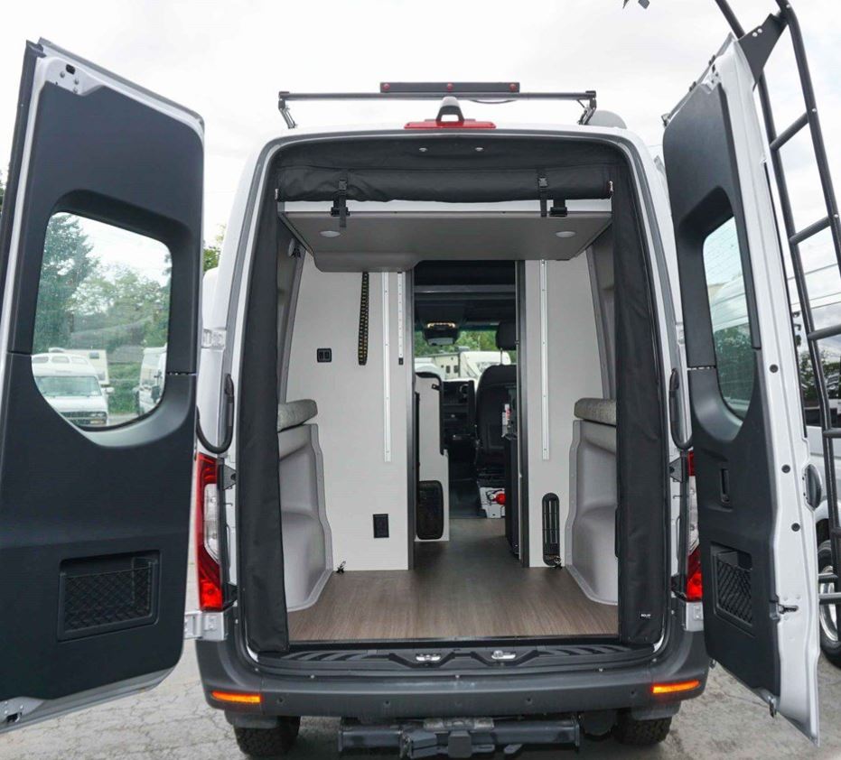 Spacious RV rental van: rear view with open doors revealing modern interior, bed platform, wood floor, & ladder on right.. Winnebago Revel 2020