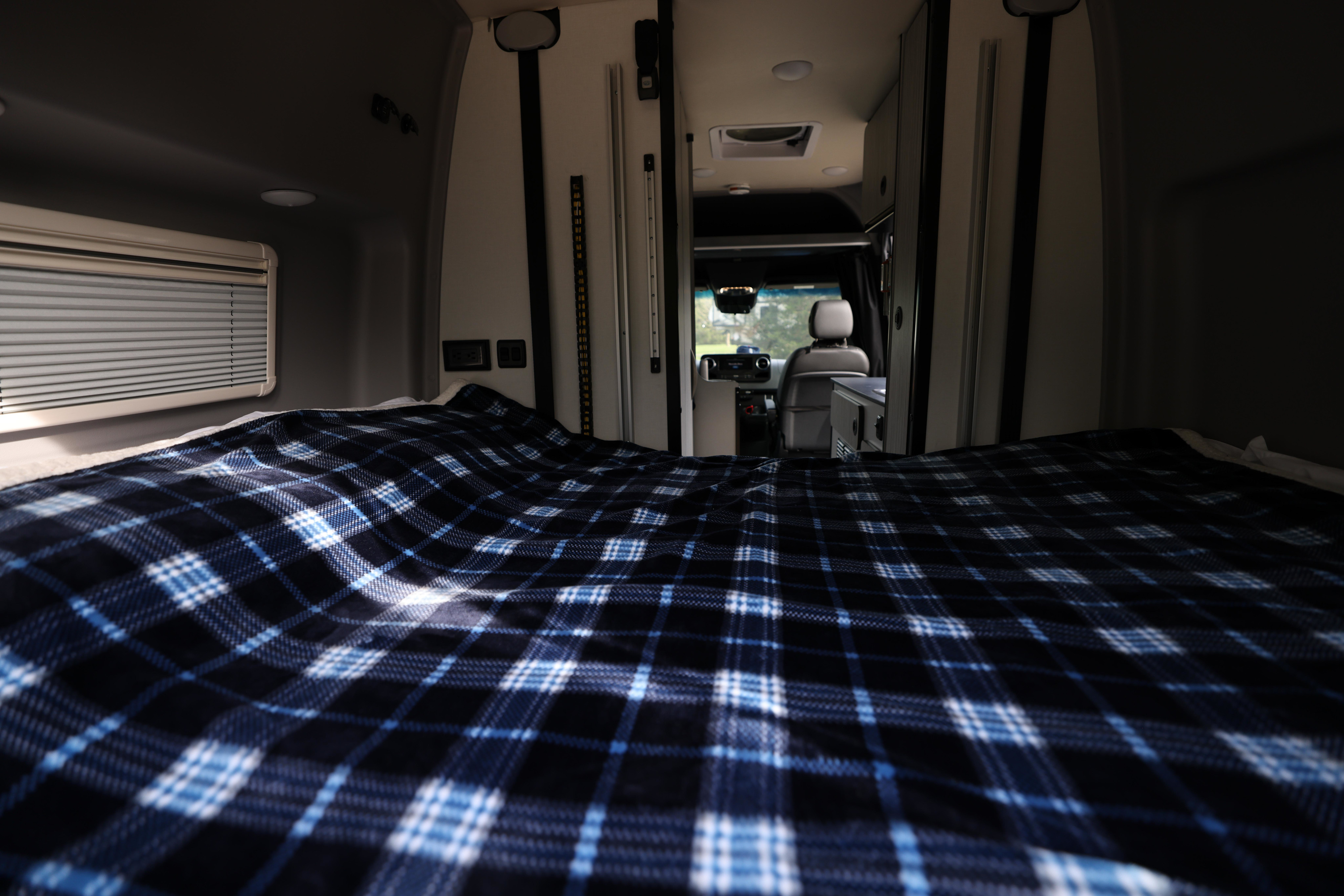 Inside an RV rental: a cozy bed with blue plaid blanket, looking forward to the driver's area. Ready for adventure.. Mercedes Winnebago Revel 2023