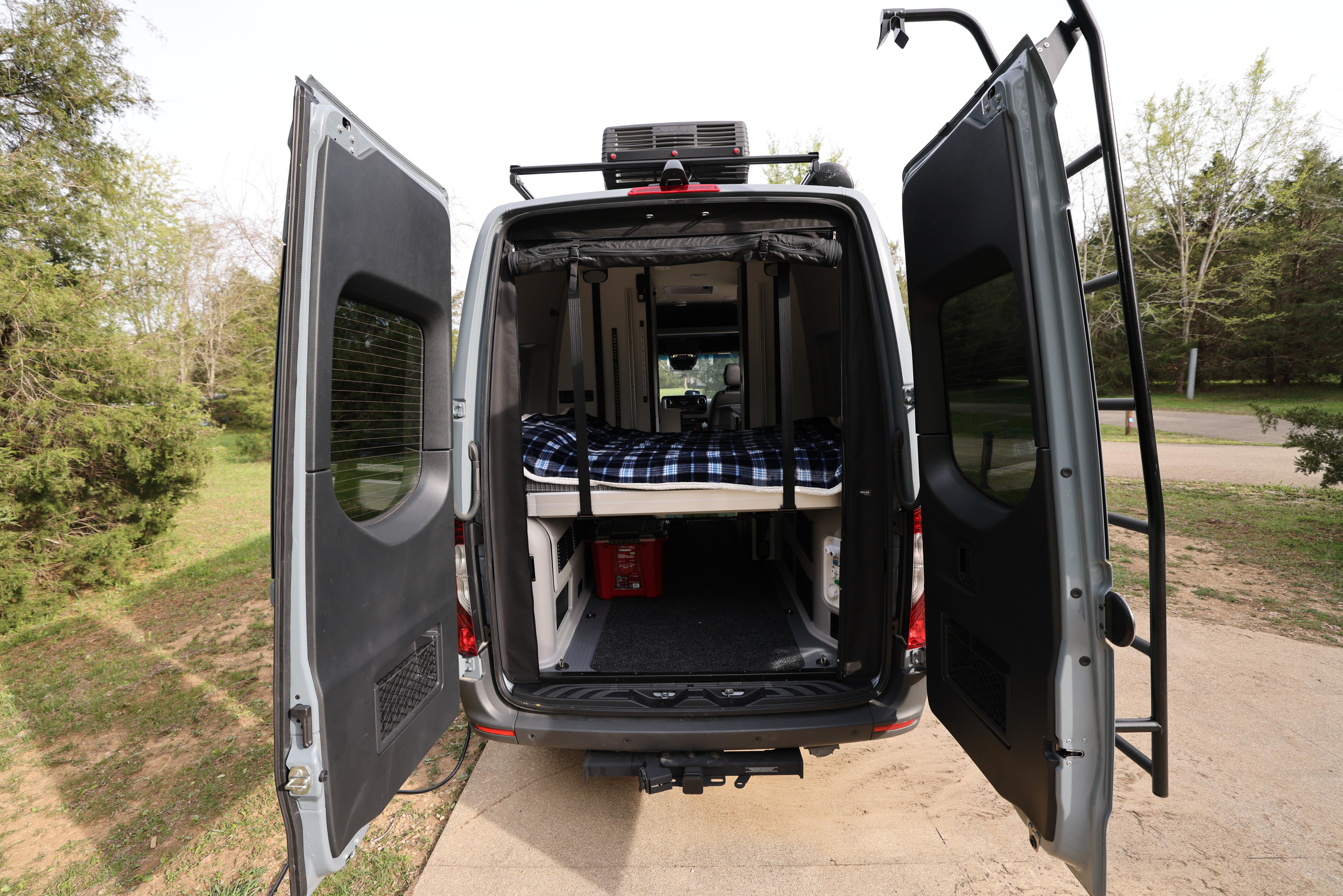 Rear view of a gray camper van RV rental, doors open revealing a comfy bed, storage, and roof access ladder.. Mercedes Winnebago Revel 2023