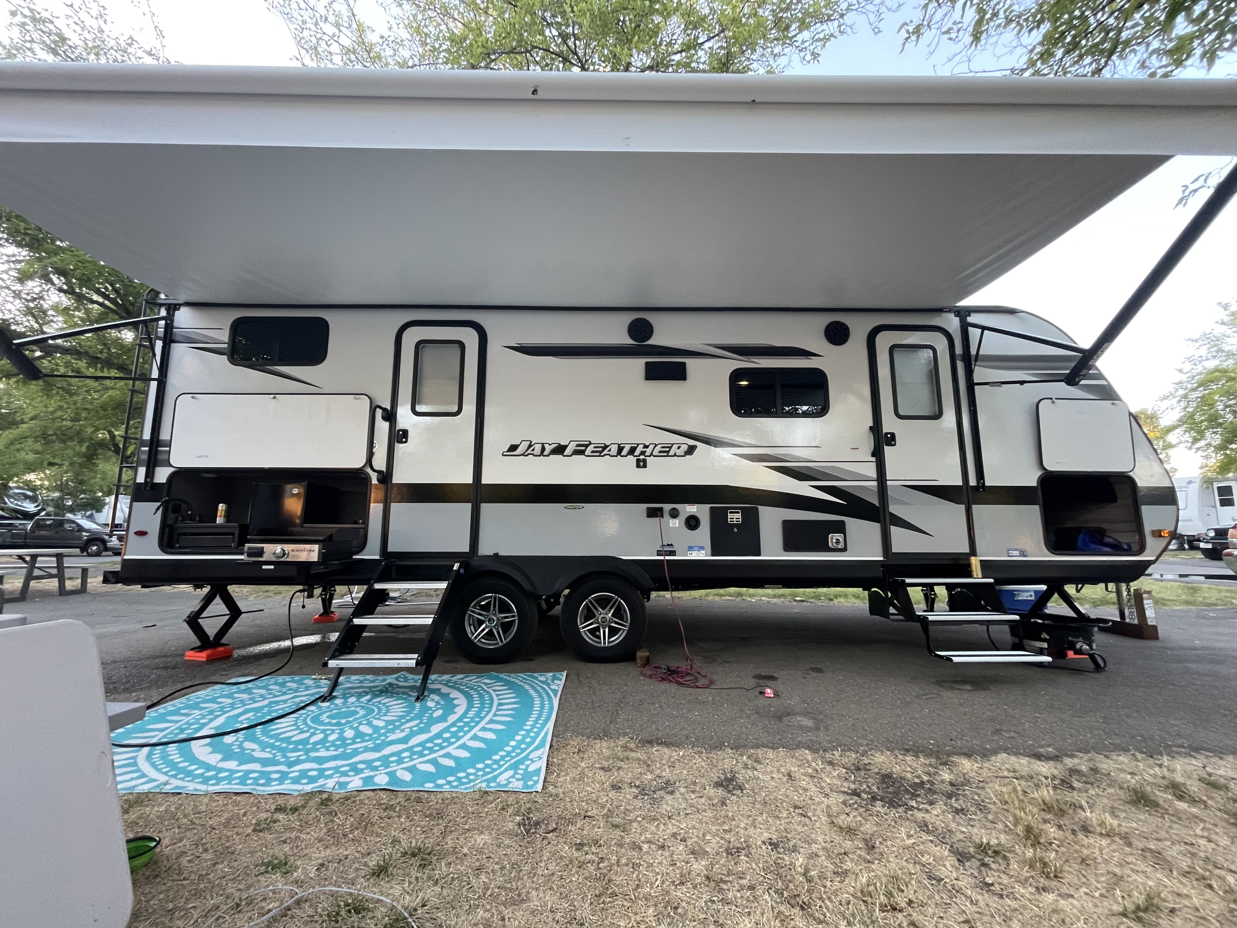 A silver Jay Feather **RV rental** sits at a **campsite**, awning extended, with an outdoor kitchen setup and a teal mat on the ground.. Jayco Jay Feather 2022