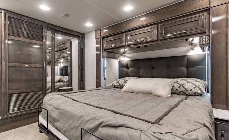 Comfortable RV bedroom interior with plush bed, sleek dark wood cabinets, and large mirror for your rv rental adventure.. Entegra Coach Reatta 2022