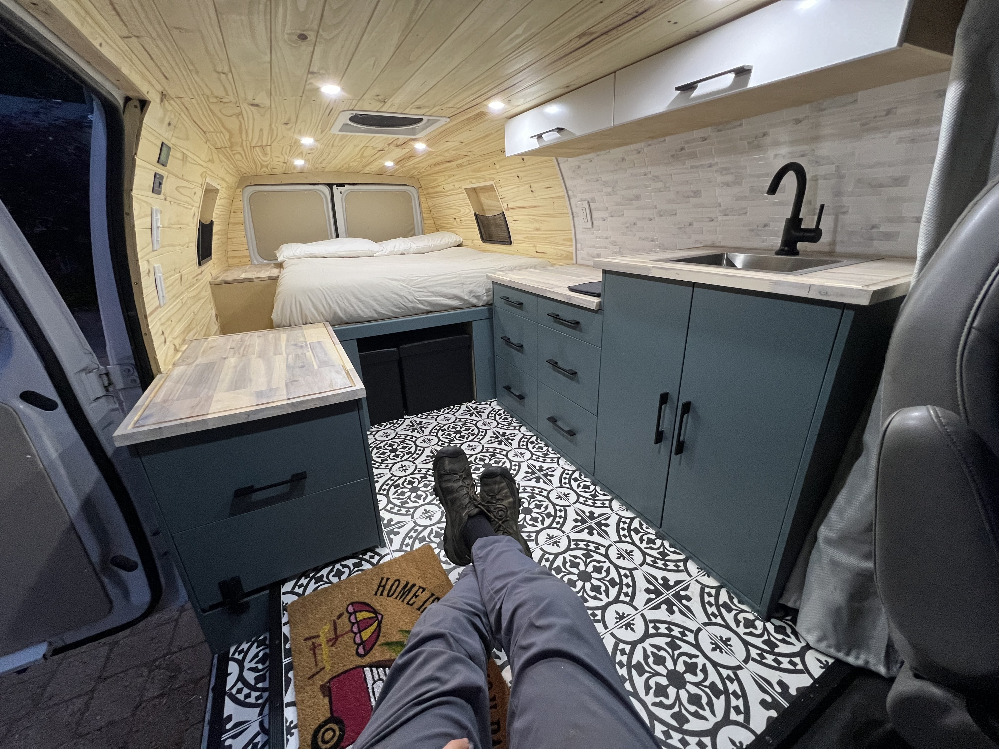 Experience the cozy interior of this RV rental, featuring a custom van build with a comfy bed, functional kitchen, and stylish patterned floor.. Ford E350 Box Van 2002