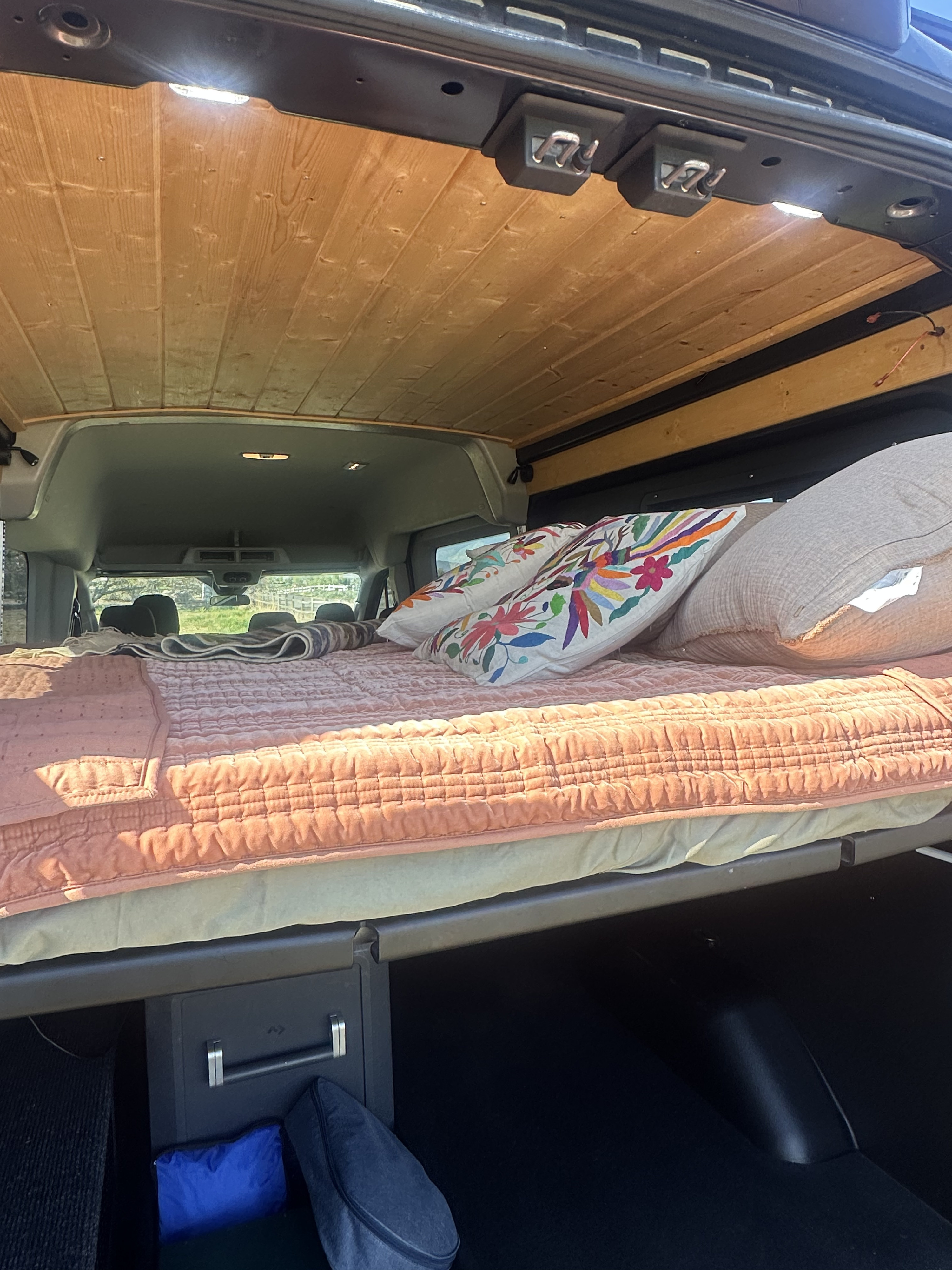 Inside an RV rental camper van: a cozy bed setup with decorative pillows, wooden ceiling, and convenient under-bed storage.. Ford Transit 2020
