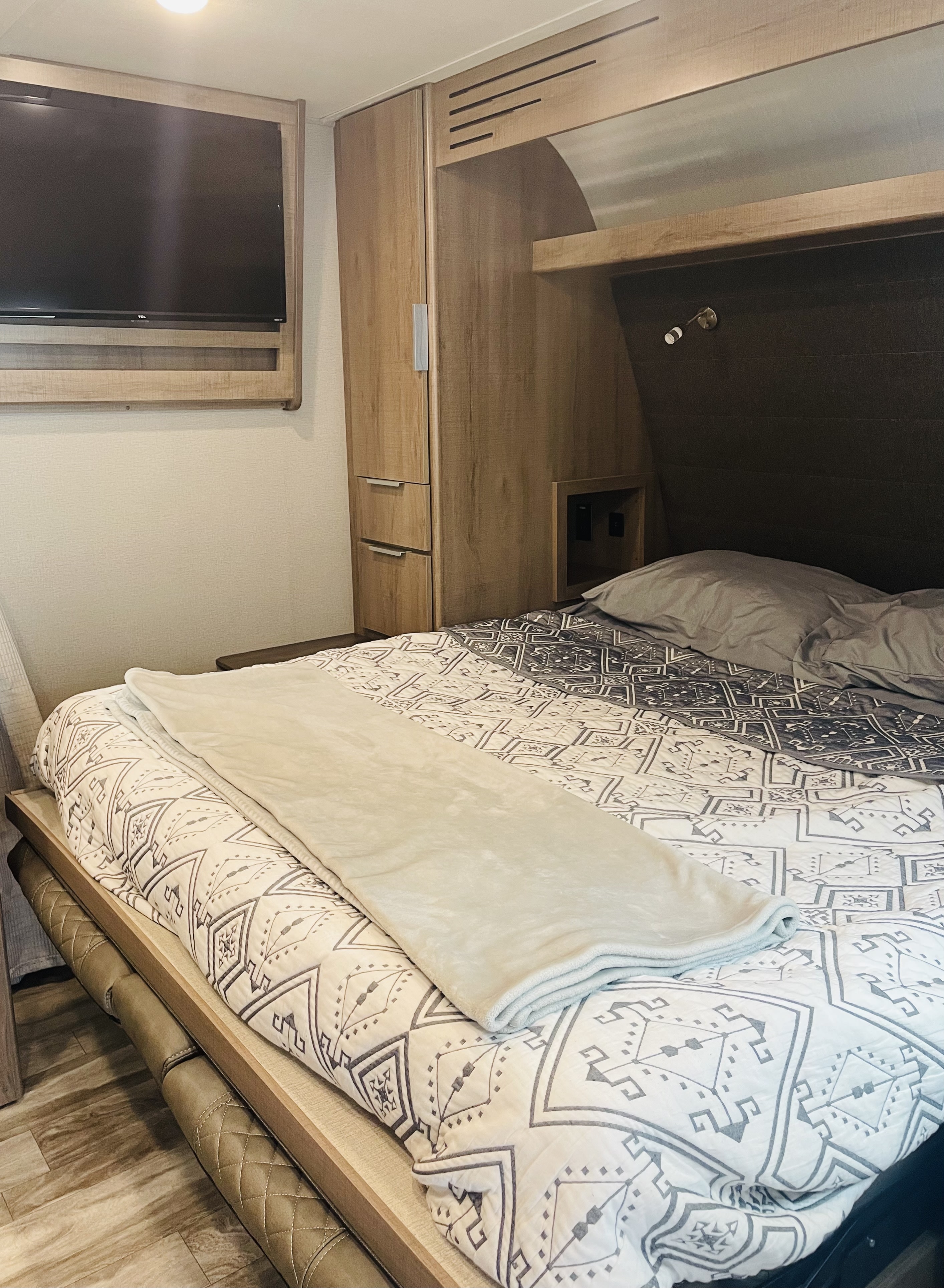 Modern RV rental bedroom, featuring a patterned bed, mounted TV, built-in storage, and reading light for comfort.. Grand Design Imagine XLS 2021