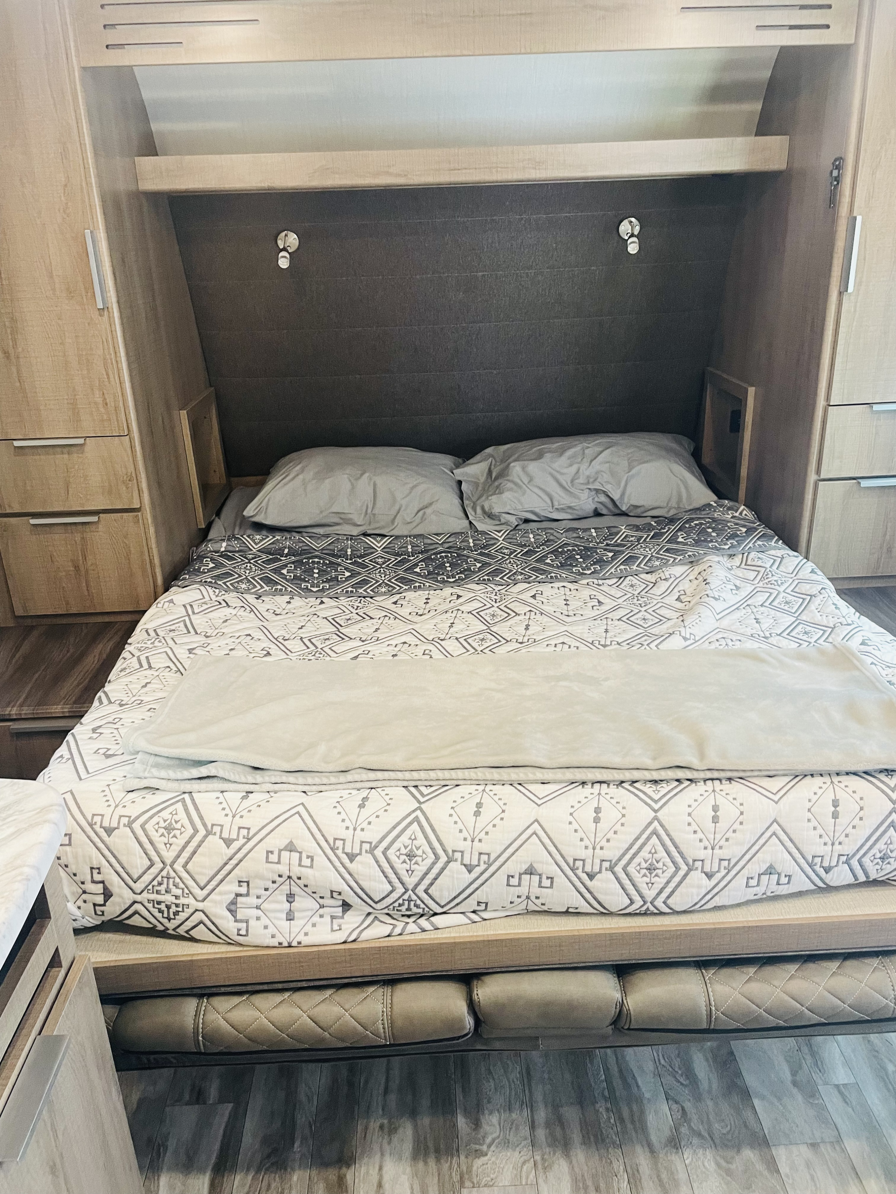 Inside an **RV rental**: a cozy bed with patterned bedding, grey pillows, and built-in wood cabinets for convenient storage.. Grand Design Imagine XLS 2021